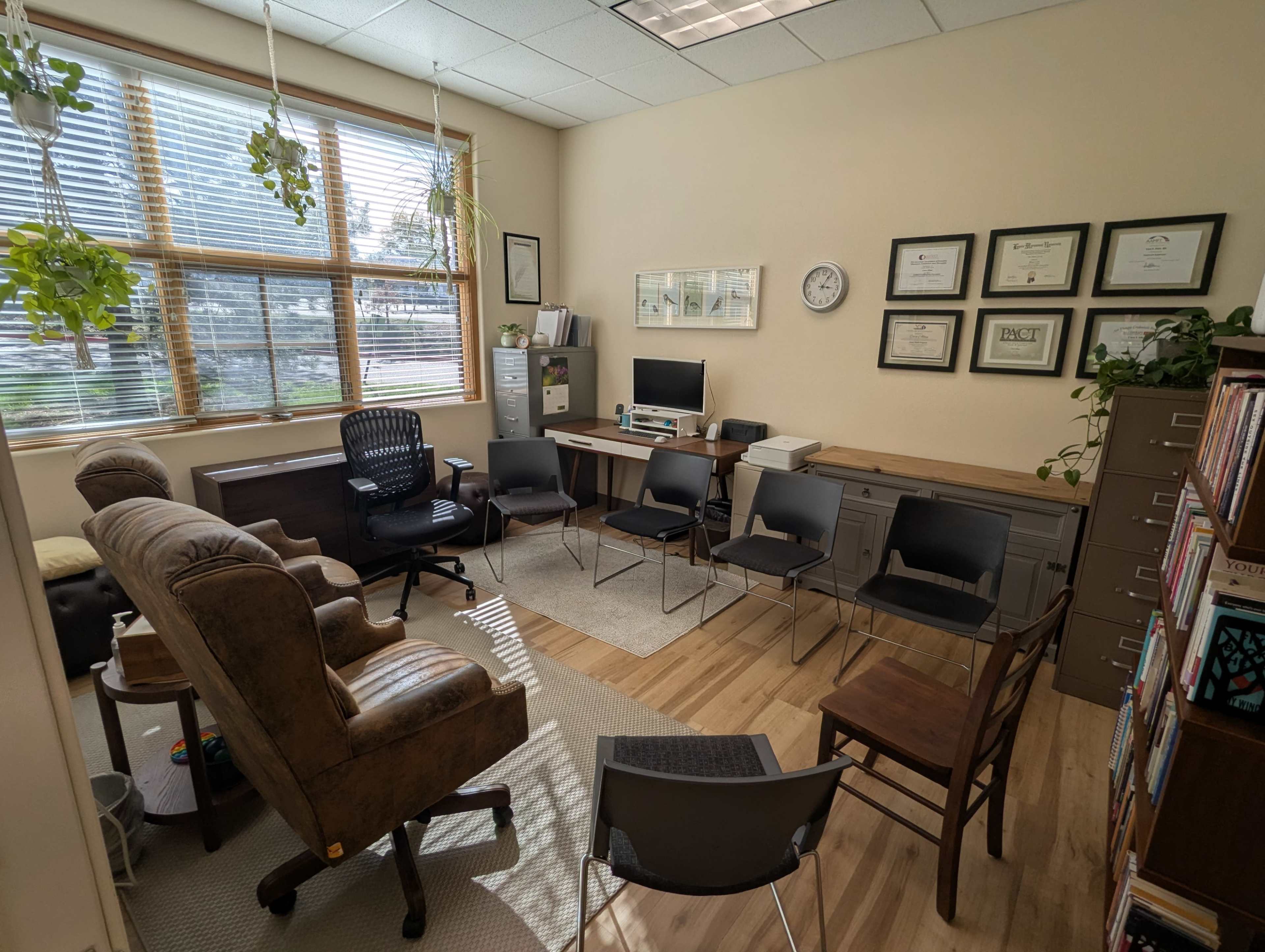 The image shows an office with a mix of seating, including two armchairs and several chairs arranged around a central space, along with a desk, computer, and filing cabinets.