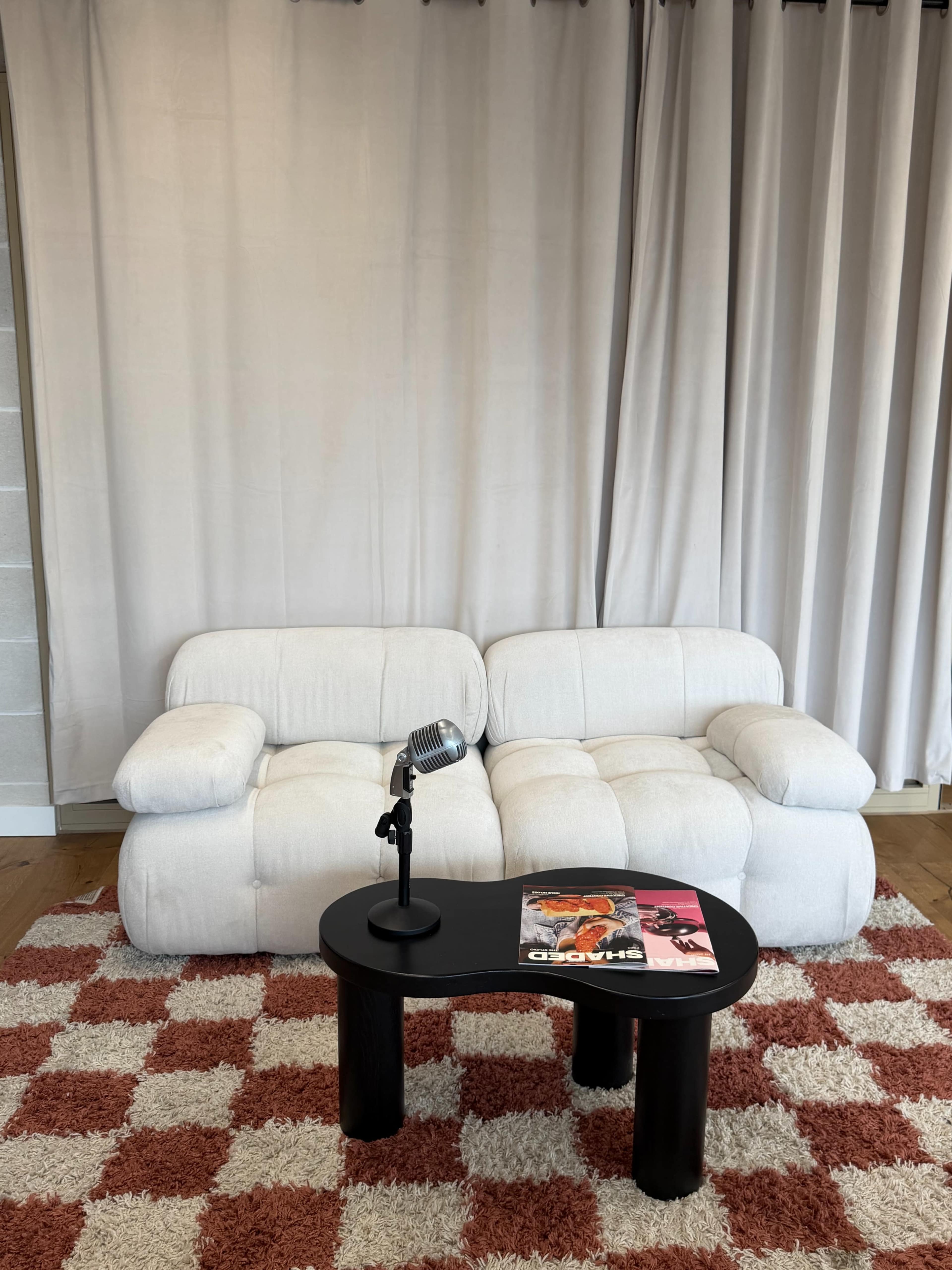 The image shows a white two-seater sofa and a small black table with a microphone and a magazine on it, set against a backdrop of light-colored curtains and a patterned rug.
