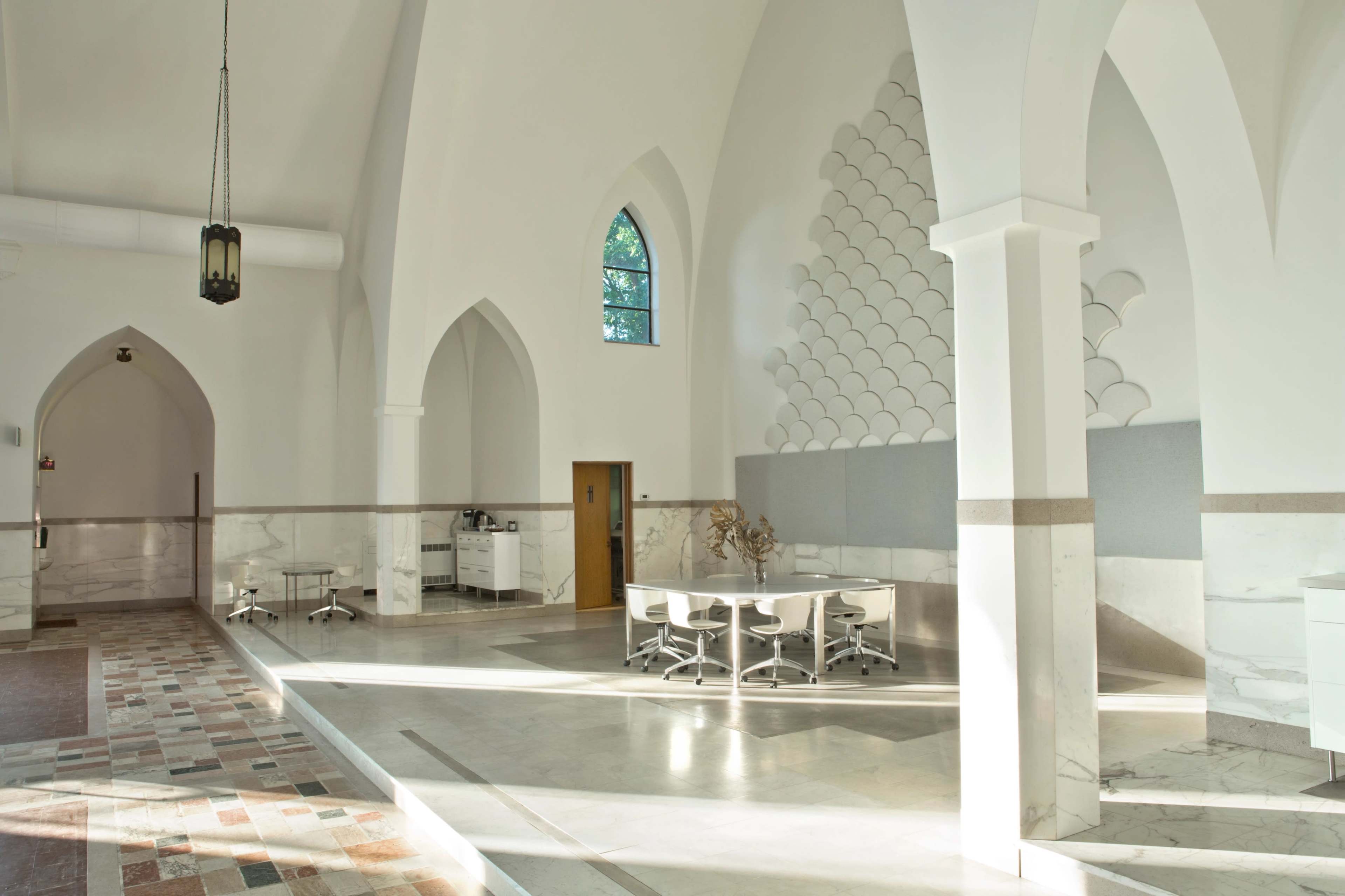 The interior of a spacious, light-filled room features high arched ceilings, tall windows, and a circular table surrounded by white chairs on a marble floor.
