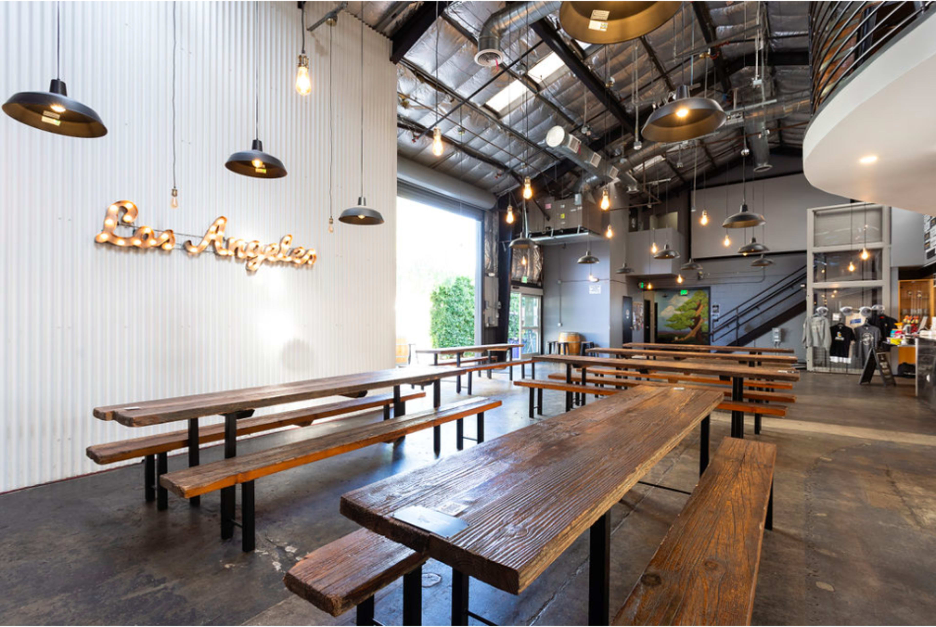 The image shows a spacious indoor area with wooden tables and benches, industrial-style lighting, and a decorative sign that reads "Los Angeles" on the wall.
