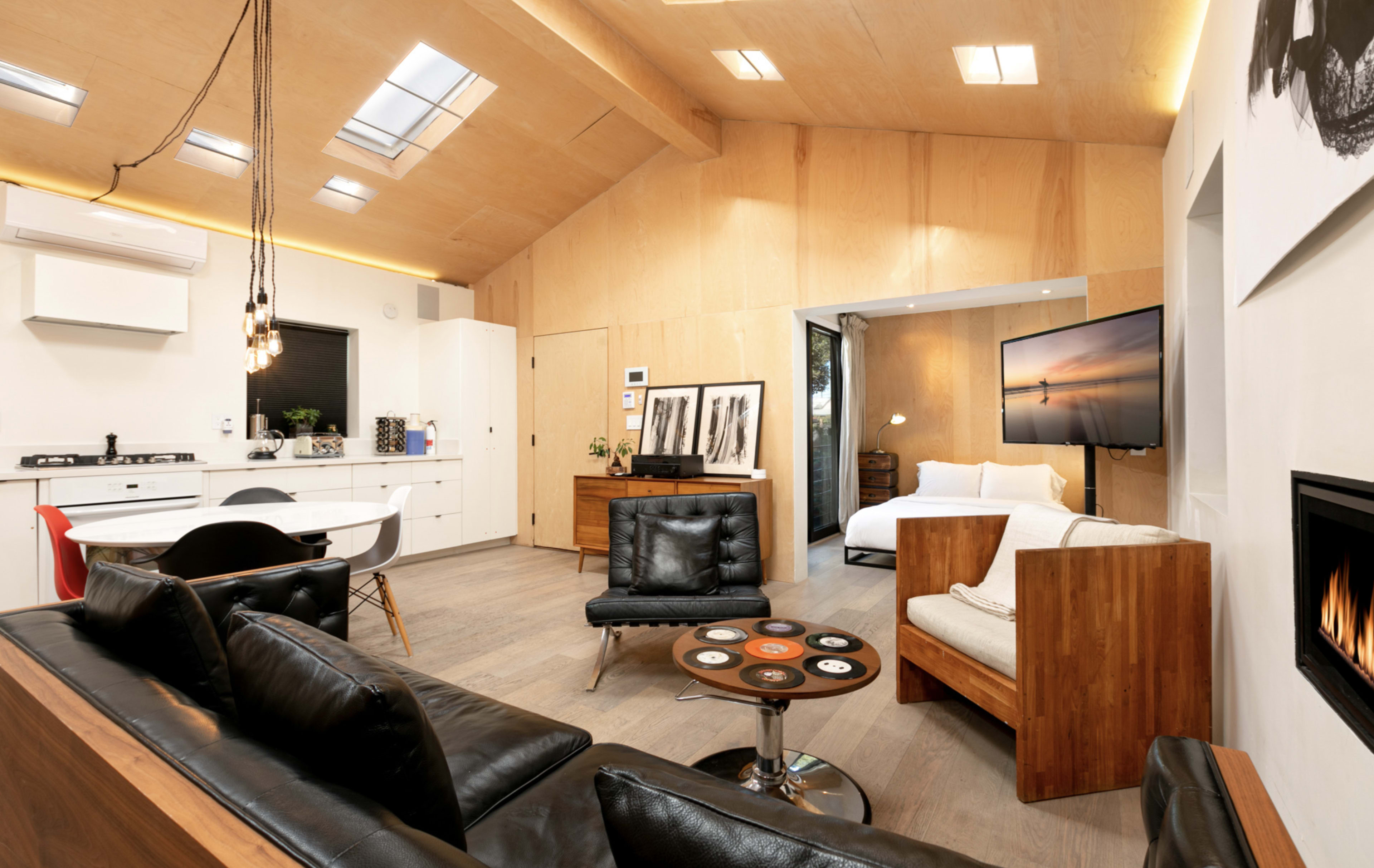 The image shows a modern, open-concept living space featuring a kitchen, a lounge area with black leather seating, and a bedroom visible through an open doorway.