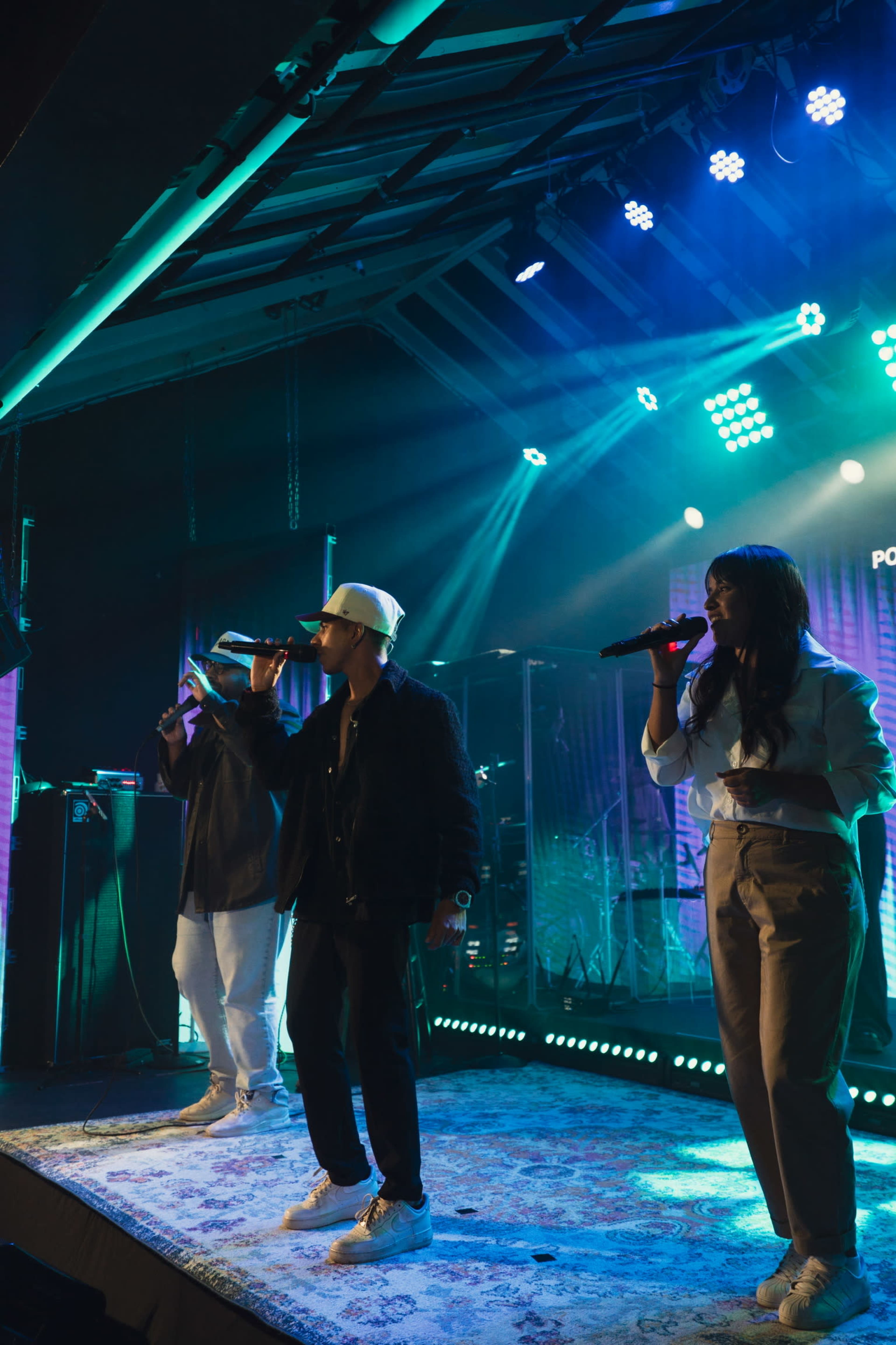Three performers are singing on stage under vibrant blue and green lights, with instruments and equipment in the background.