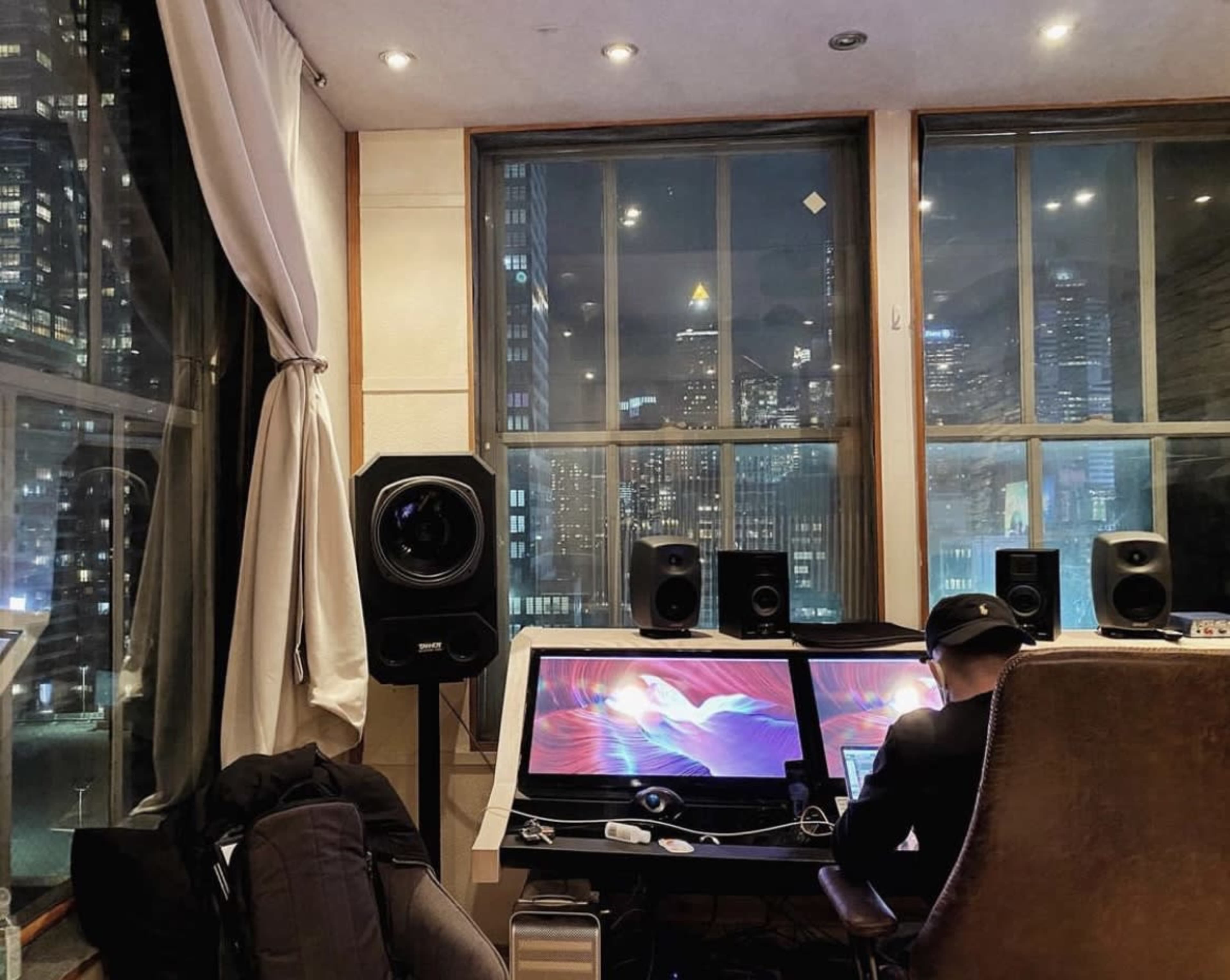 A person sits in a chair working at a desk with two computer monitors in a room with large windows overlooking a city skyline at night.