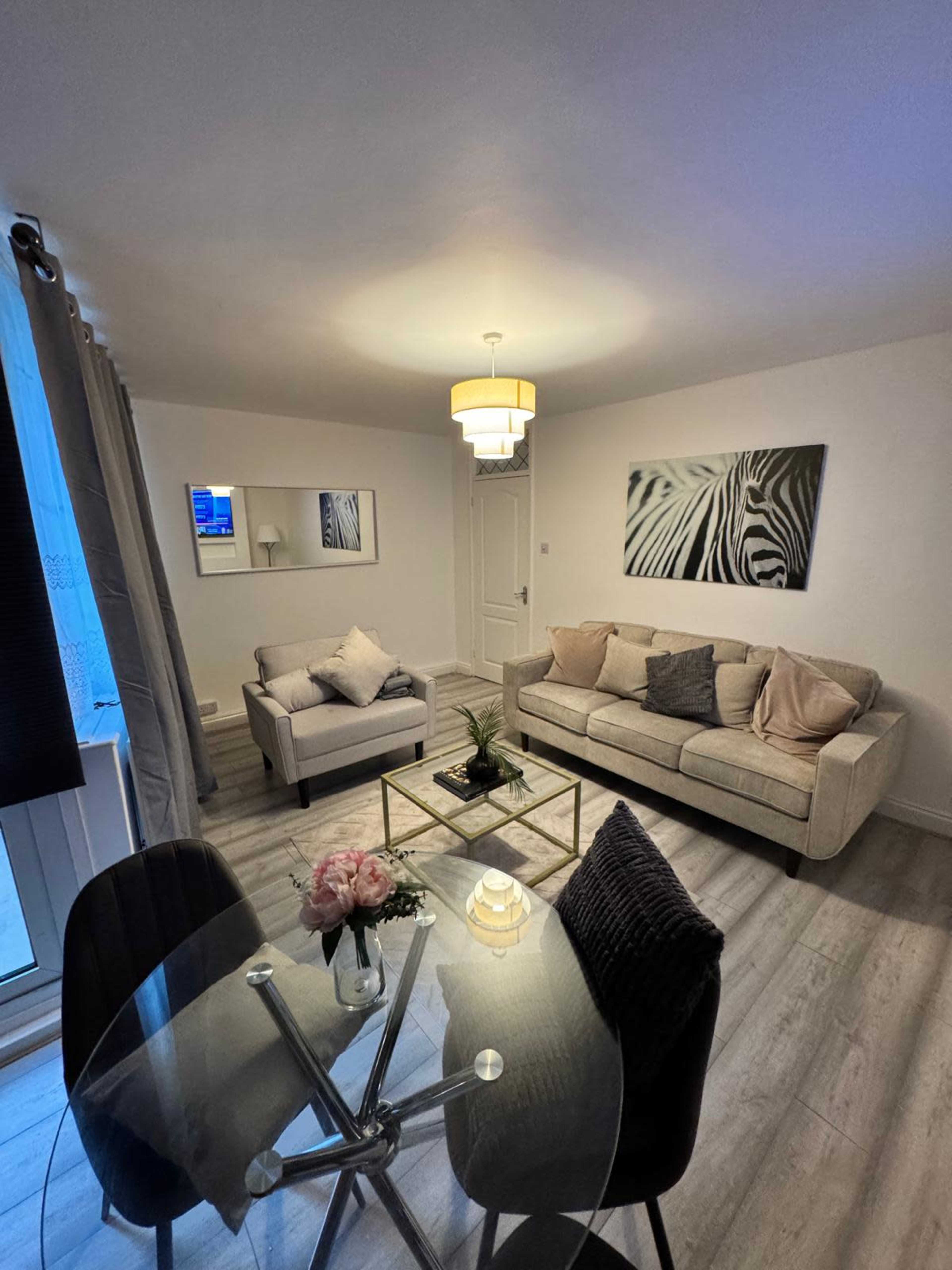The image shows a modern living room with a beige sofa, a matching loveseat, a glass coffee table, and a zebra-striped wall art piece, illuminated by a ceiling light.