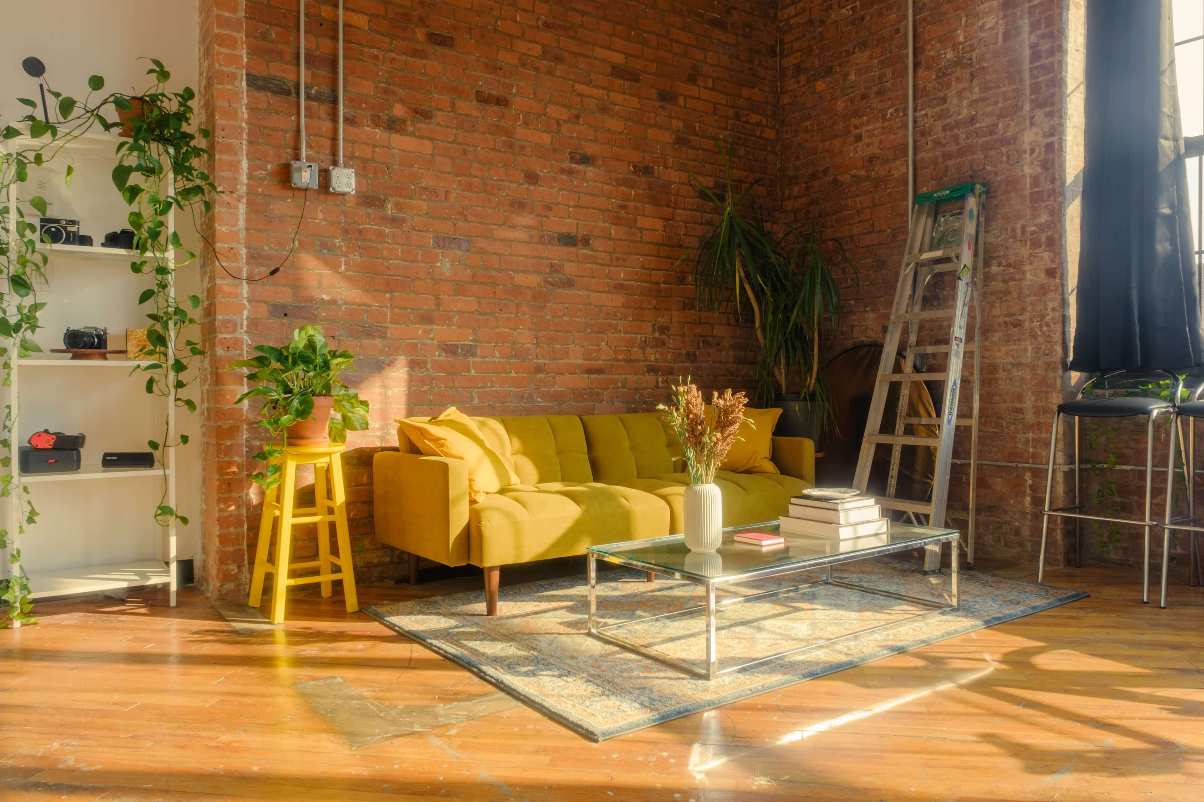 The image shows a cozy living space featuring a yellow sofa, a glass coffee table, a ladder, potted plants, and a brick wall backdrop.