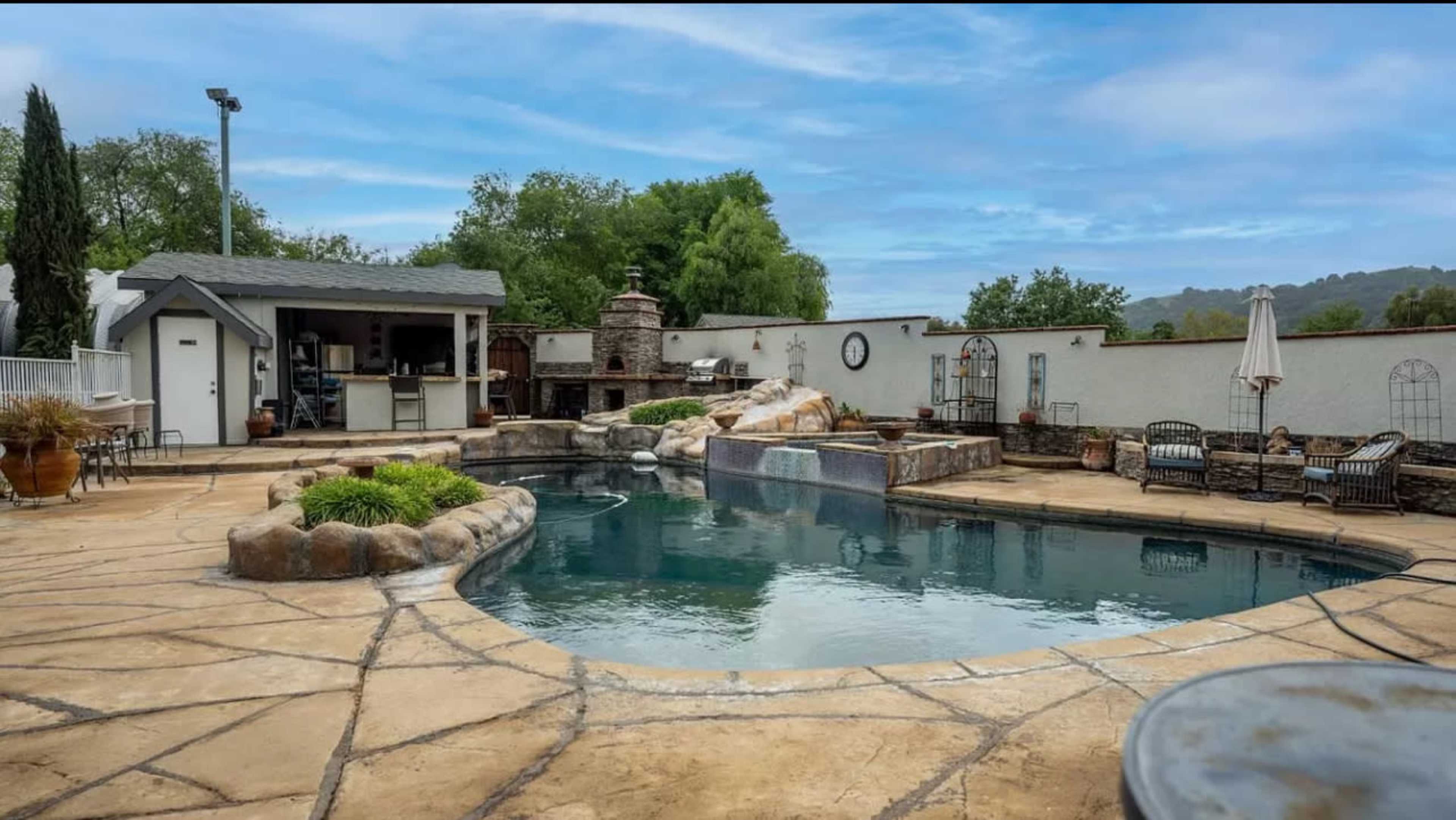 A landscaped backyard featuring a rectangular swimming pool surrounded by stone decking, a small waterfall, and seating areas with a barbecue station in the background.
