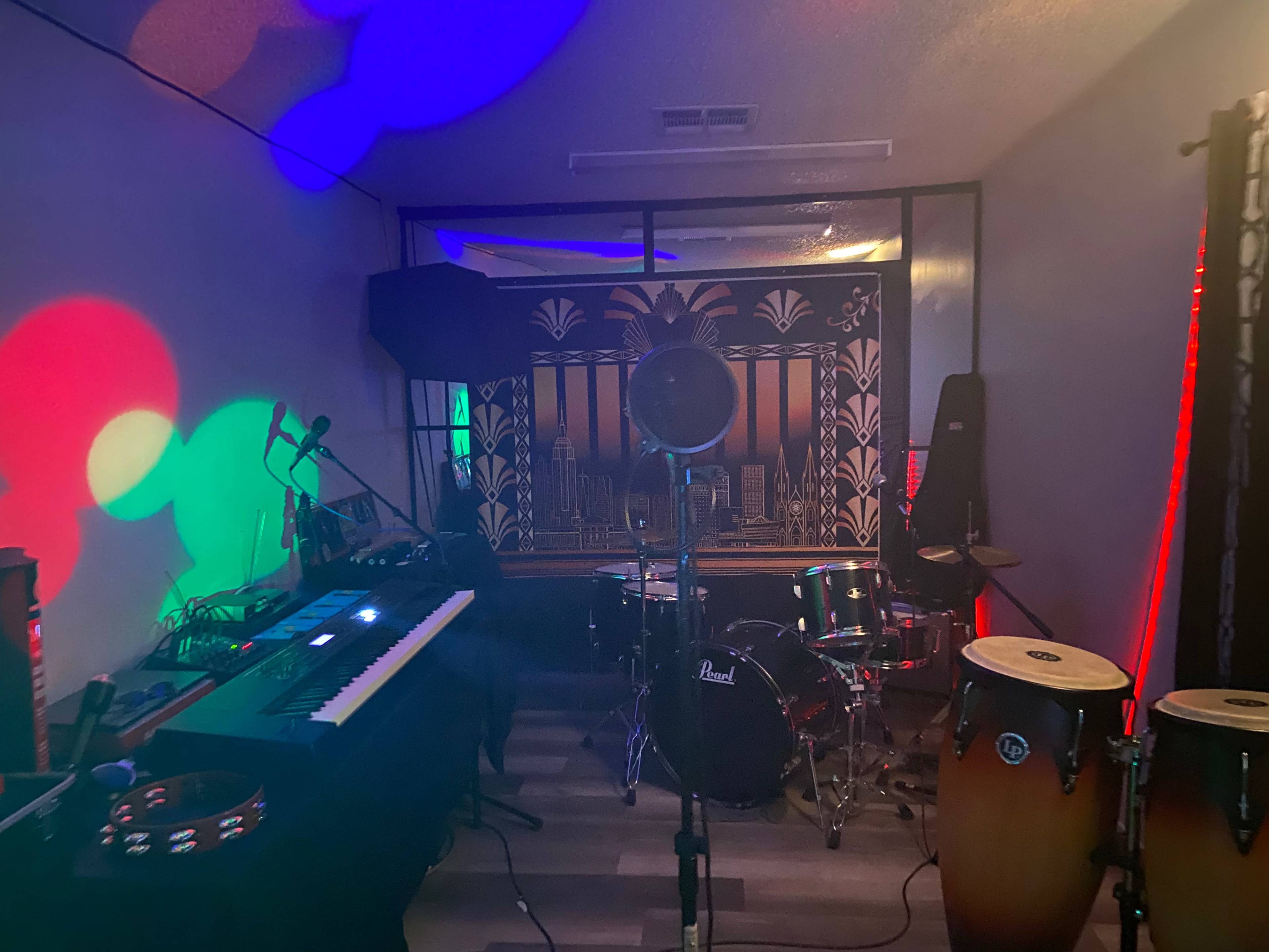 A music studio setup featuring a keyboard, drums, conga drums, and colorful lighting effects on the walls.