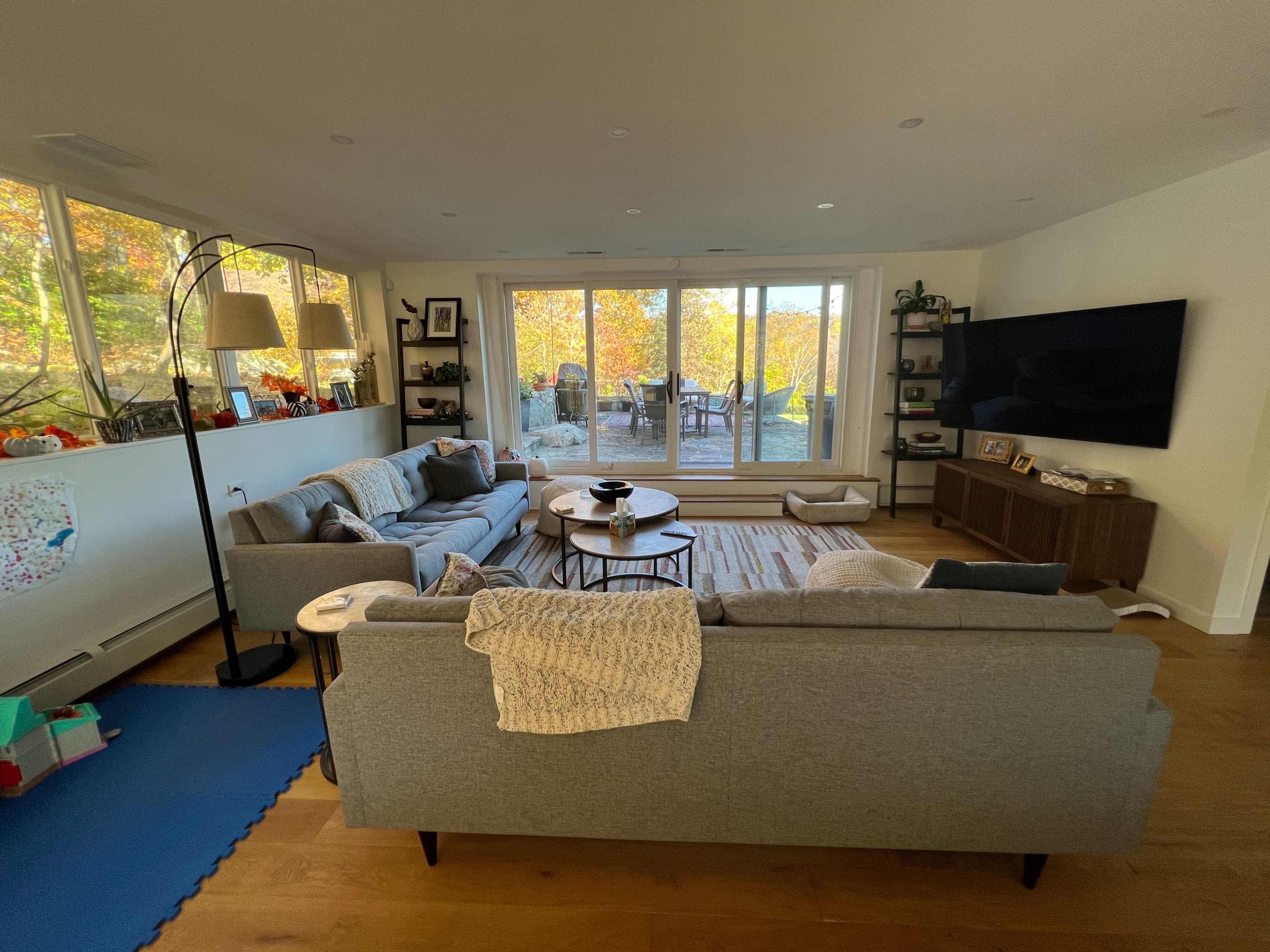 A modern living room features two gray sofas facing a large window with views of an outdoor patio and autumn foliage.