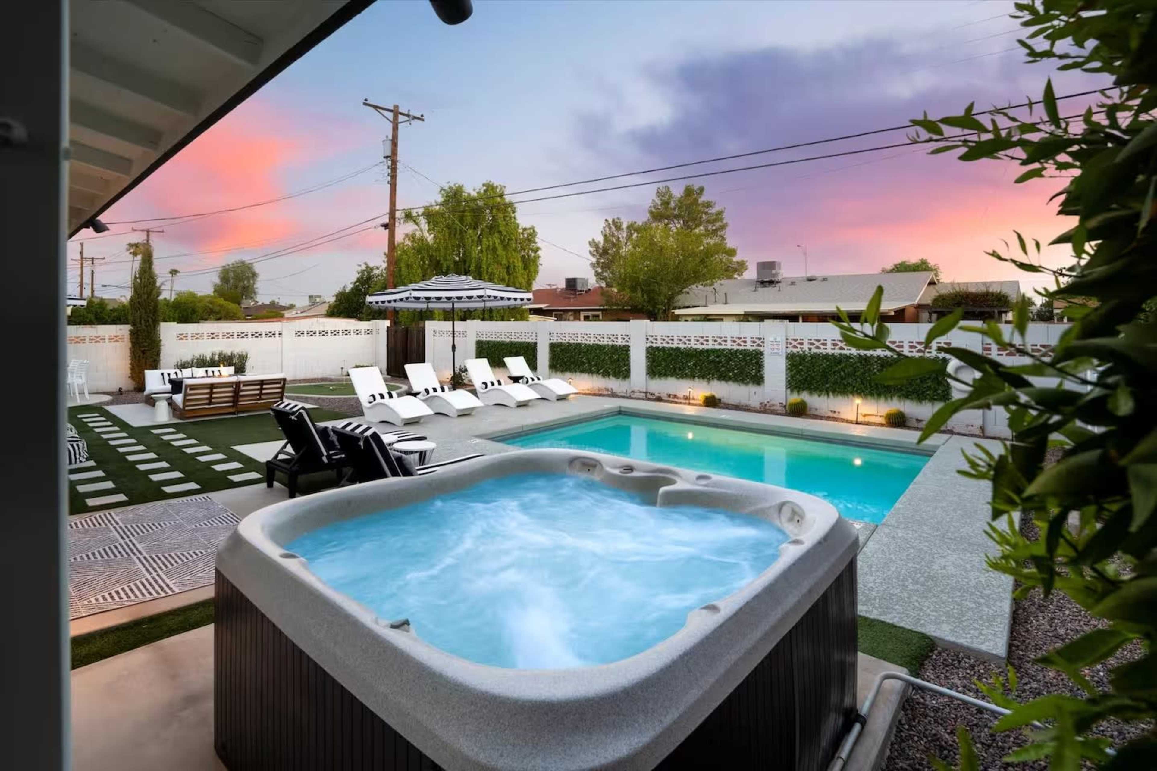A hot tub is located next to a swimming pool in a backyard with lounge chairs and a covered patio under a vibrant sky at dusk.