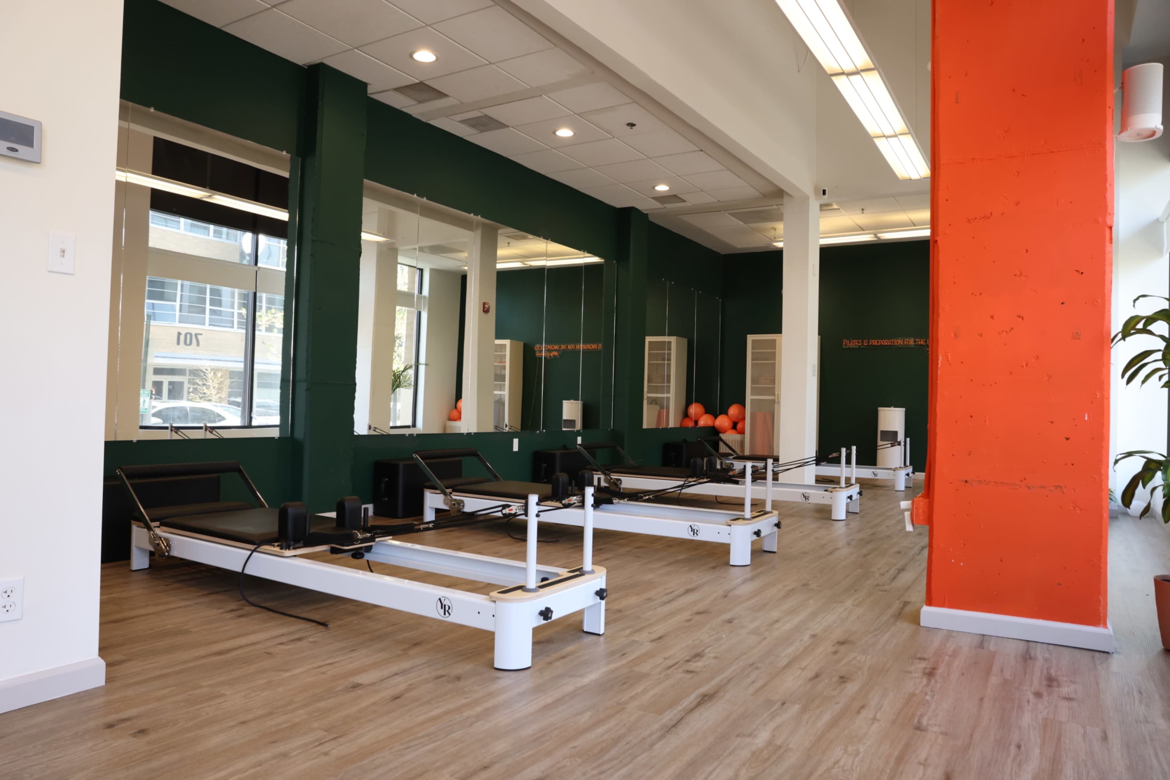 The image shows a fitness studio with several pilates reformer machines arranged in a neatly organized space with green and orange walls.
