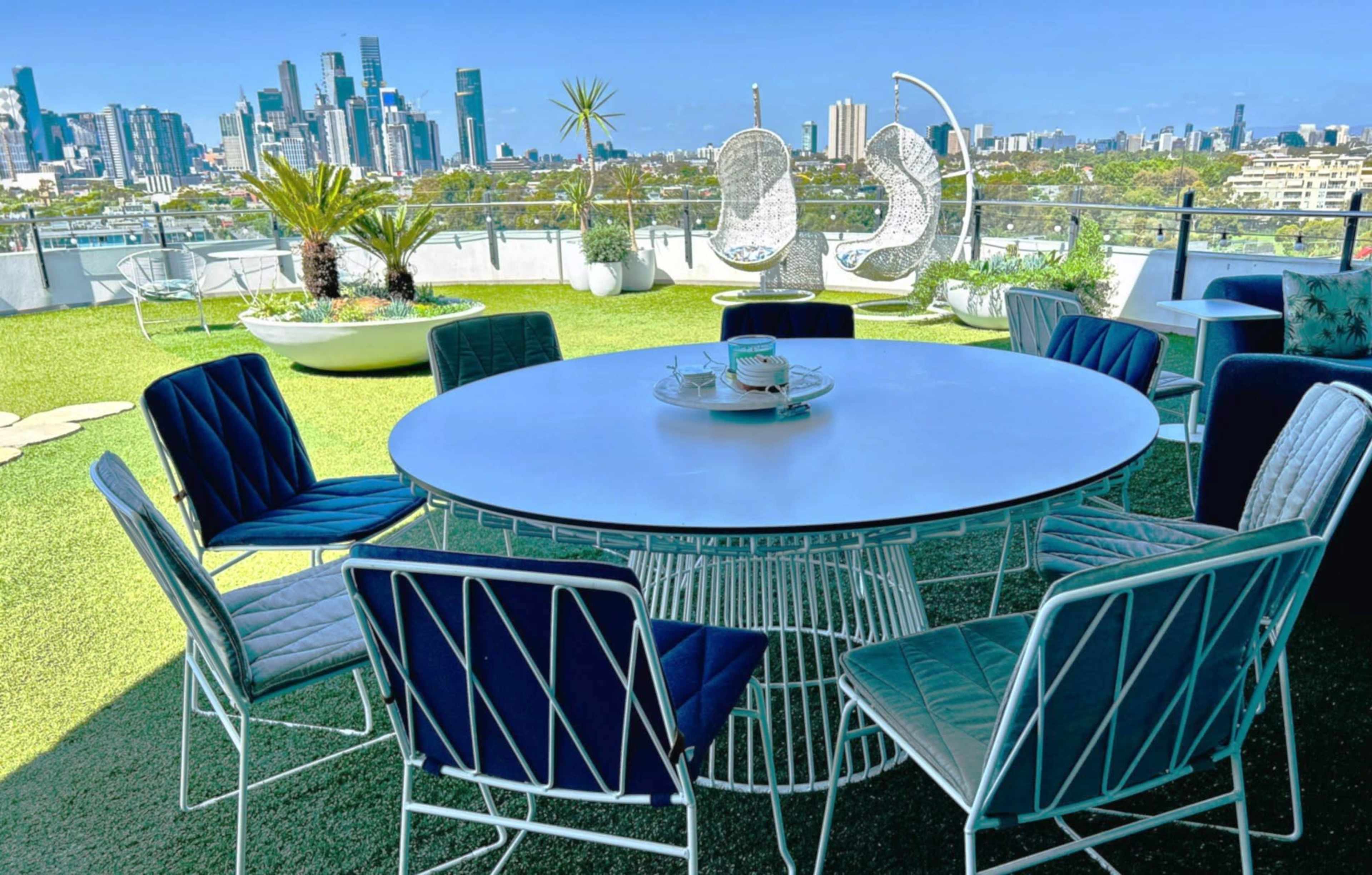 The image shows a circular table surrounded by eight chairs on a rooftop terrace with city skyline views in the background.