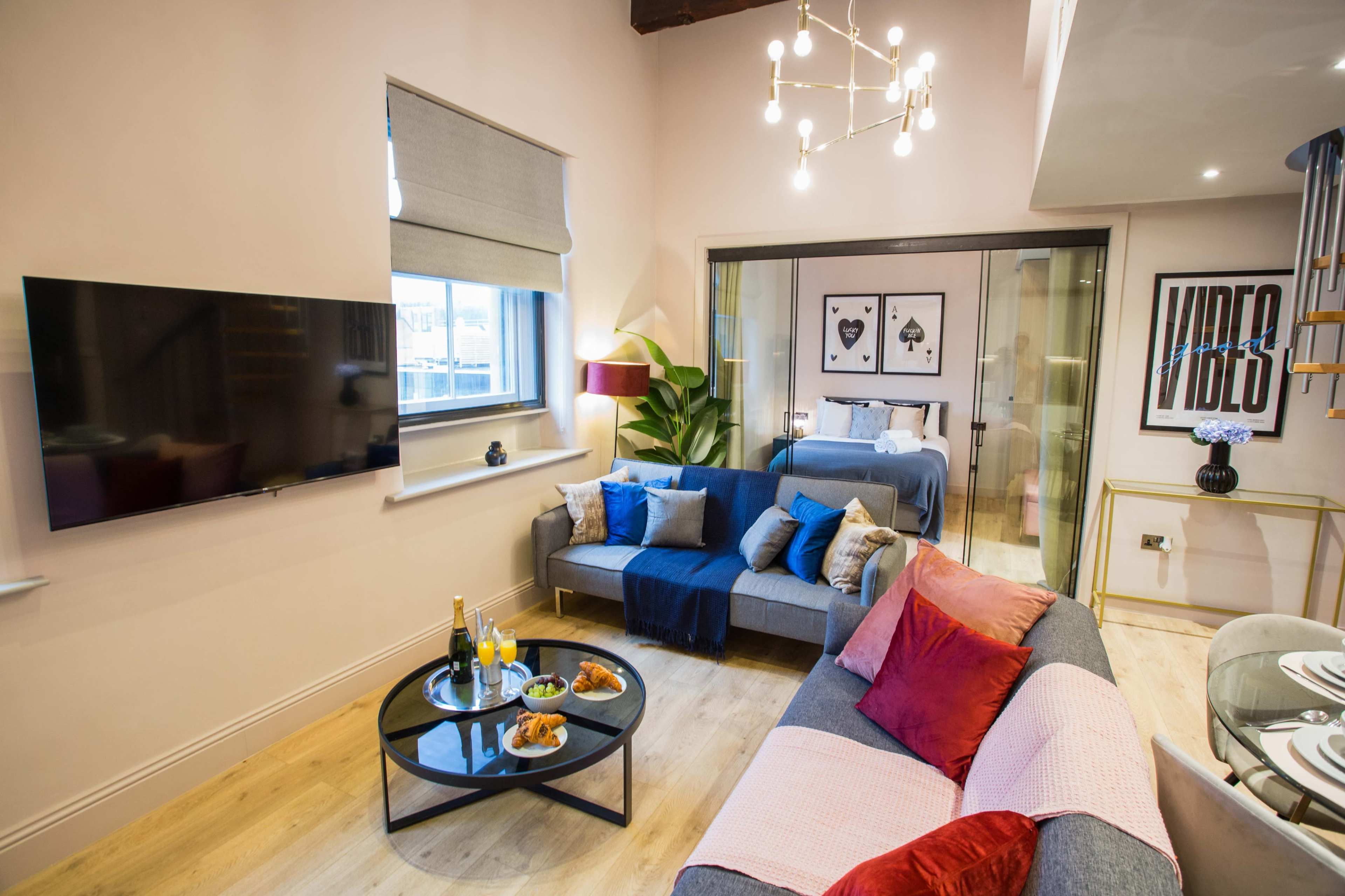 The image shows a modern living room with a television mounted on the wall, a coffee table with food and drinks, and comfortable seating arranged around a light-colored floor.