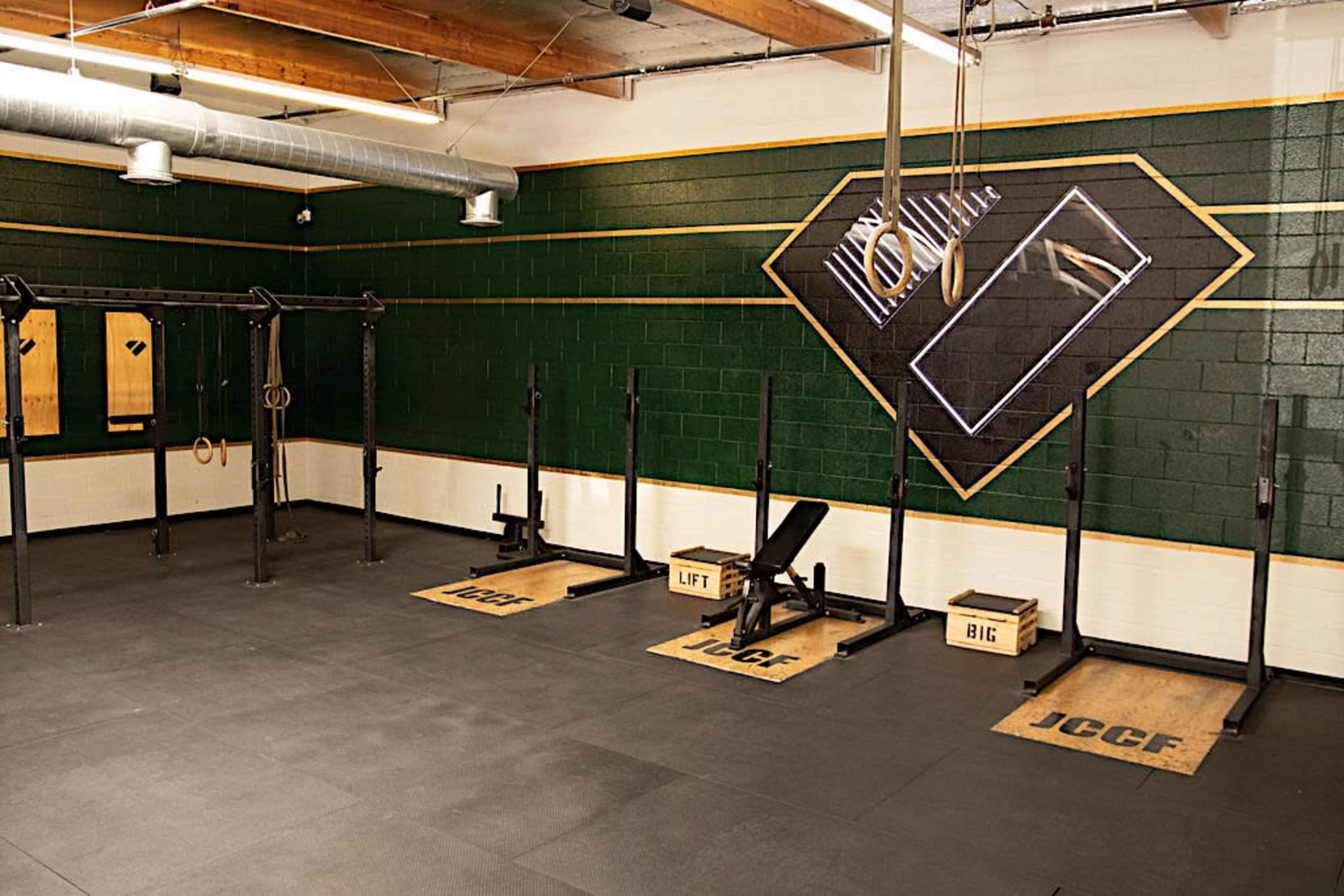 A gym with dark green walls, featuring multiple weightlifting racks and mats labeled "JCCF," "LIFT," and "BIG."