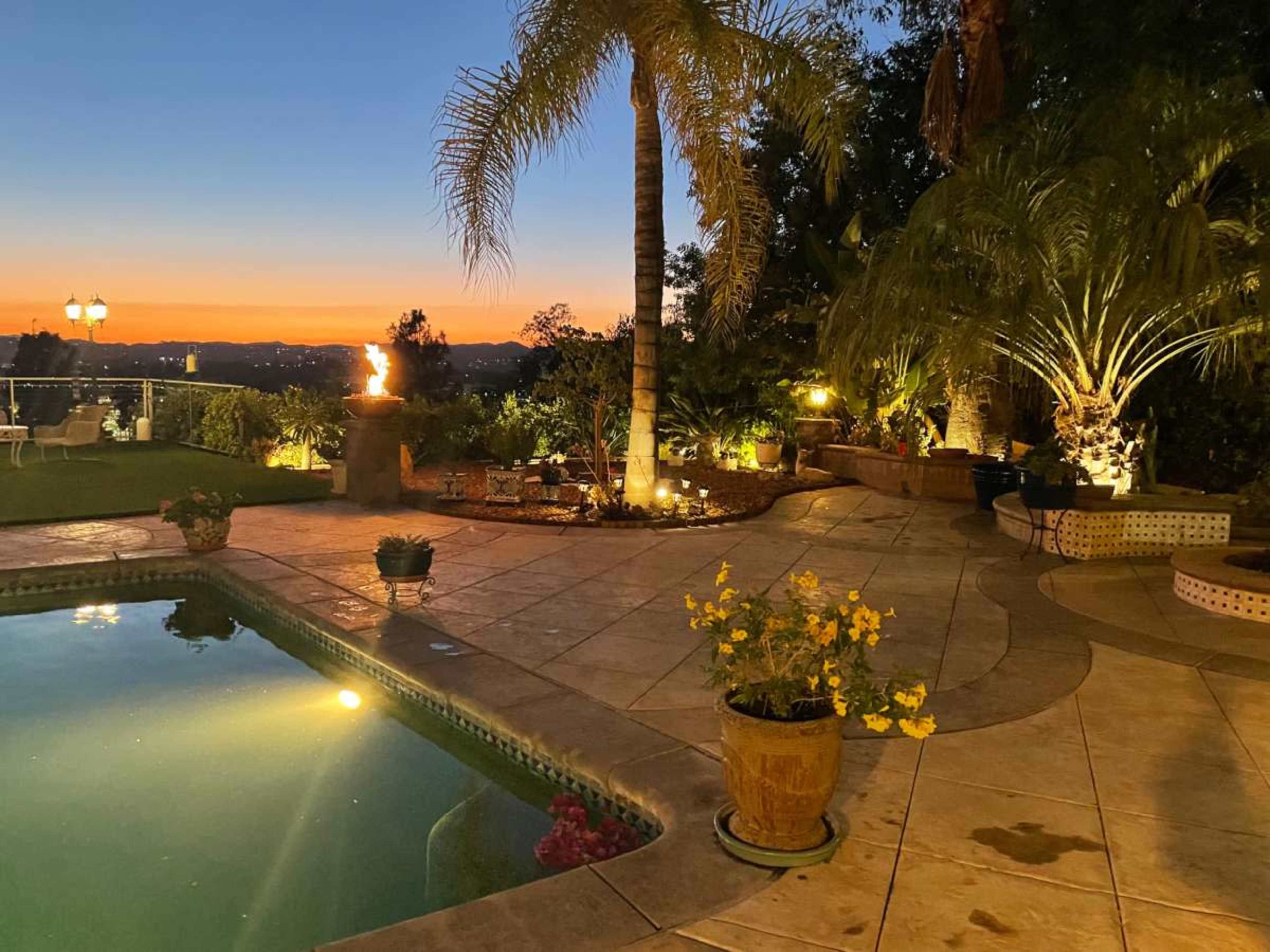 The scene features a landscaped patio with a swimming pool, palm trees, and flower pots, set against a sunset backdrop over distant hills.