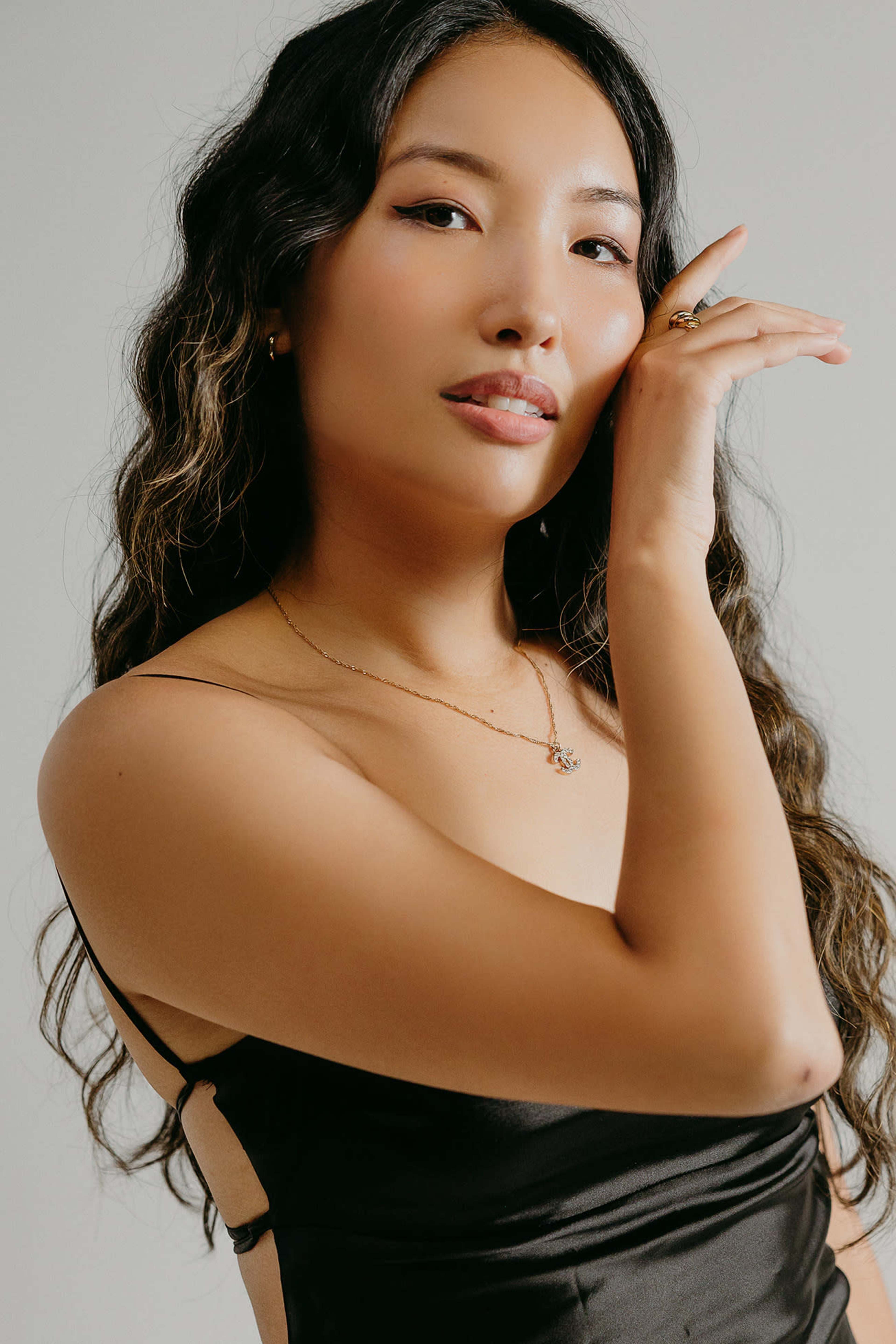 A woman with wavy hair poses while gently touching her face, wearing a black spaghetti strap dress and simple jewelry.