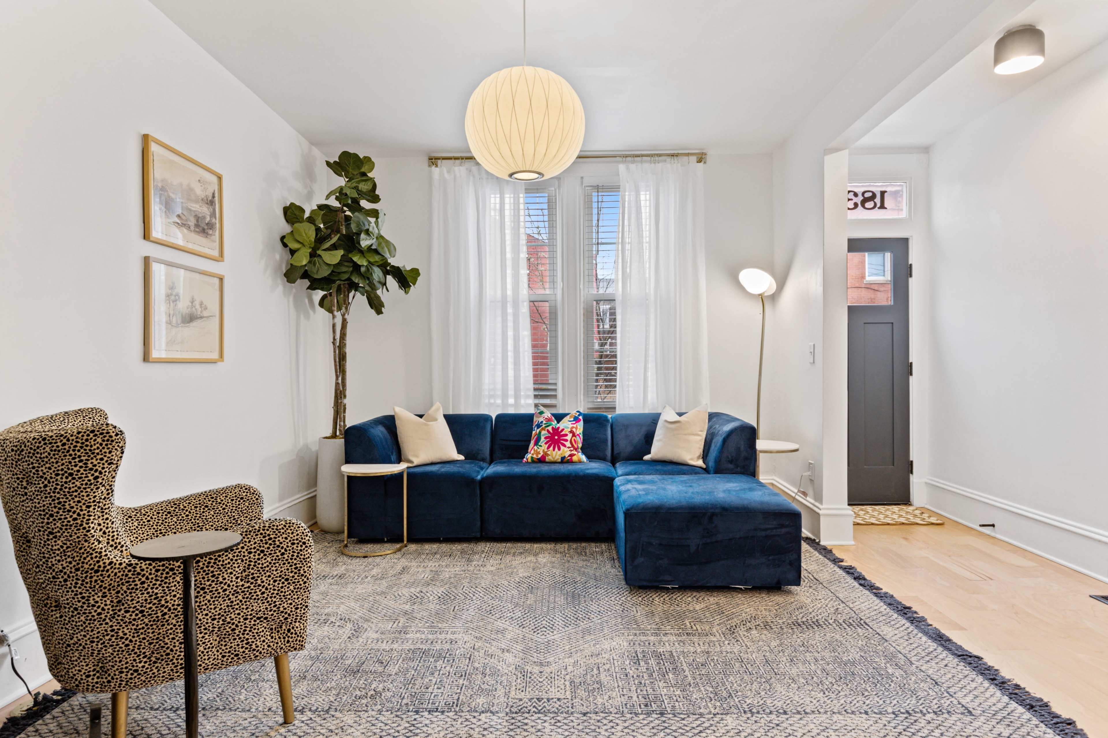 The living room features a blue sectional sofa, a patterned chair, a floor plant, and a round pendant light, complemented by a rug and framed artwork on the walls.