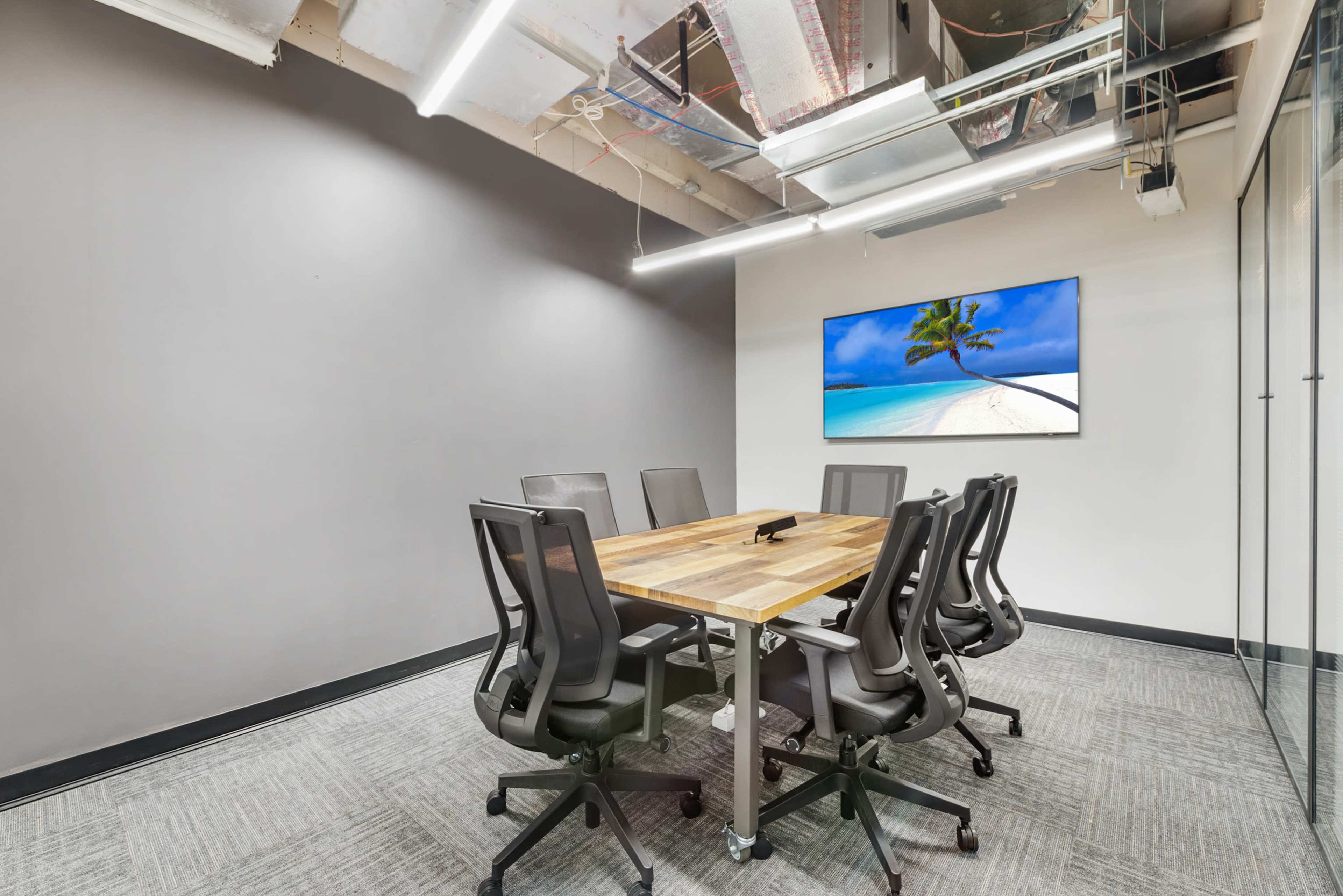 A modern conference room features a large wooden table surrounded by ergonomic chairs, with a wall-mounted television displaying a beach scene.