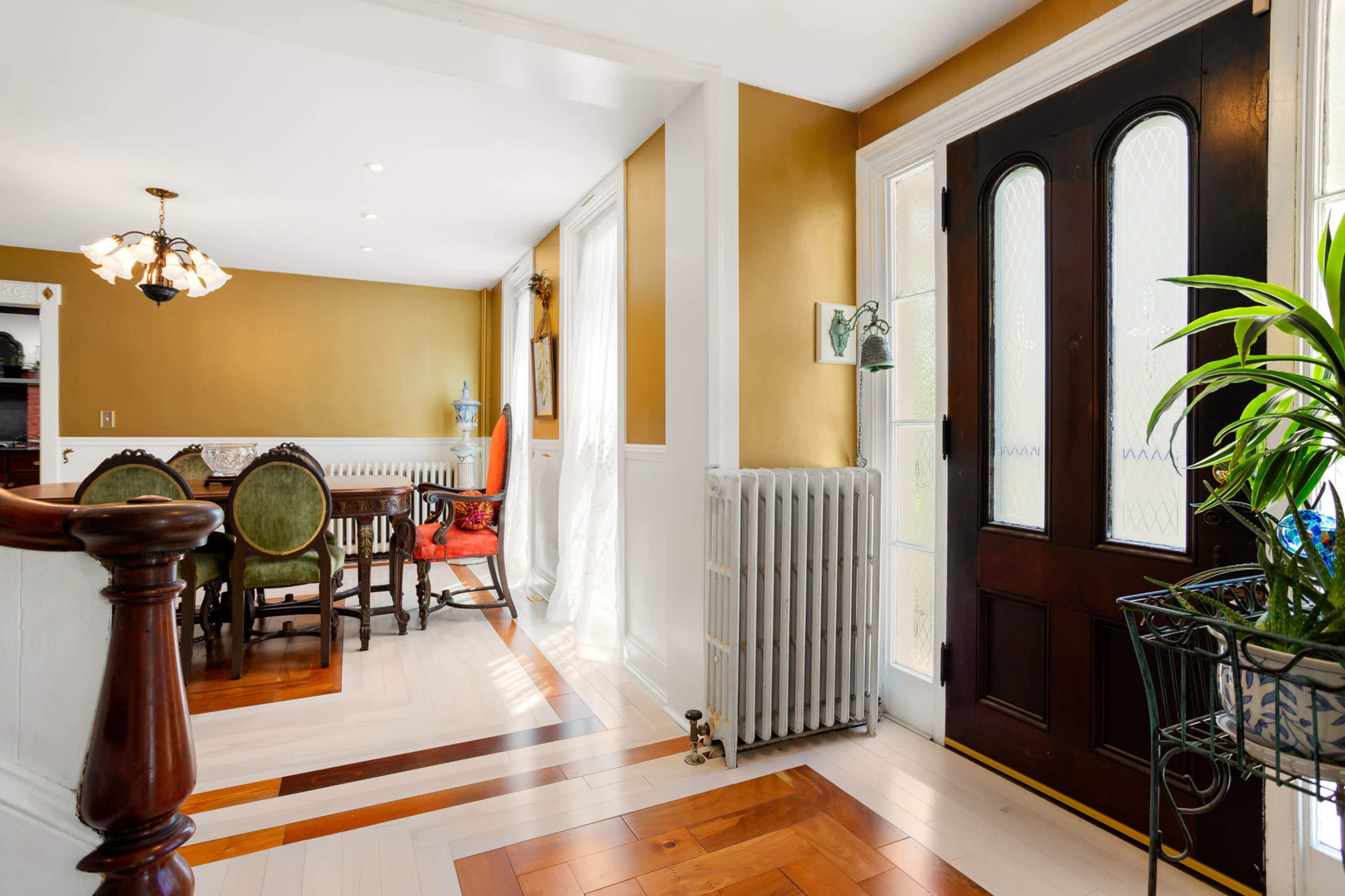 A well-lit entryway leading to a dining area with a wooden table, surrounded by chairs, and features a decorative radiator and a plant in a pot.