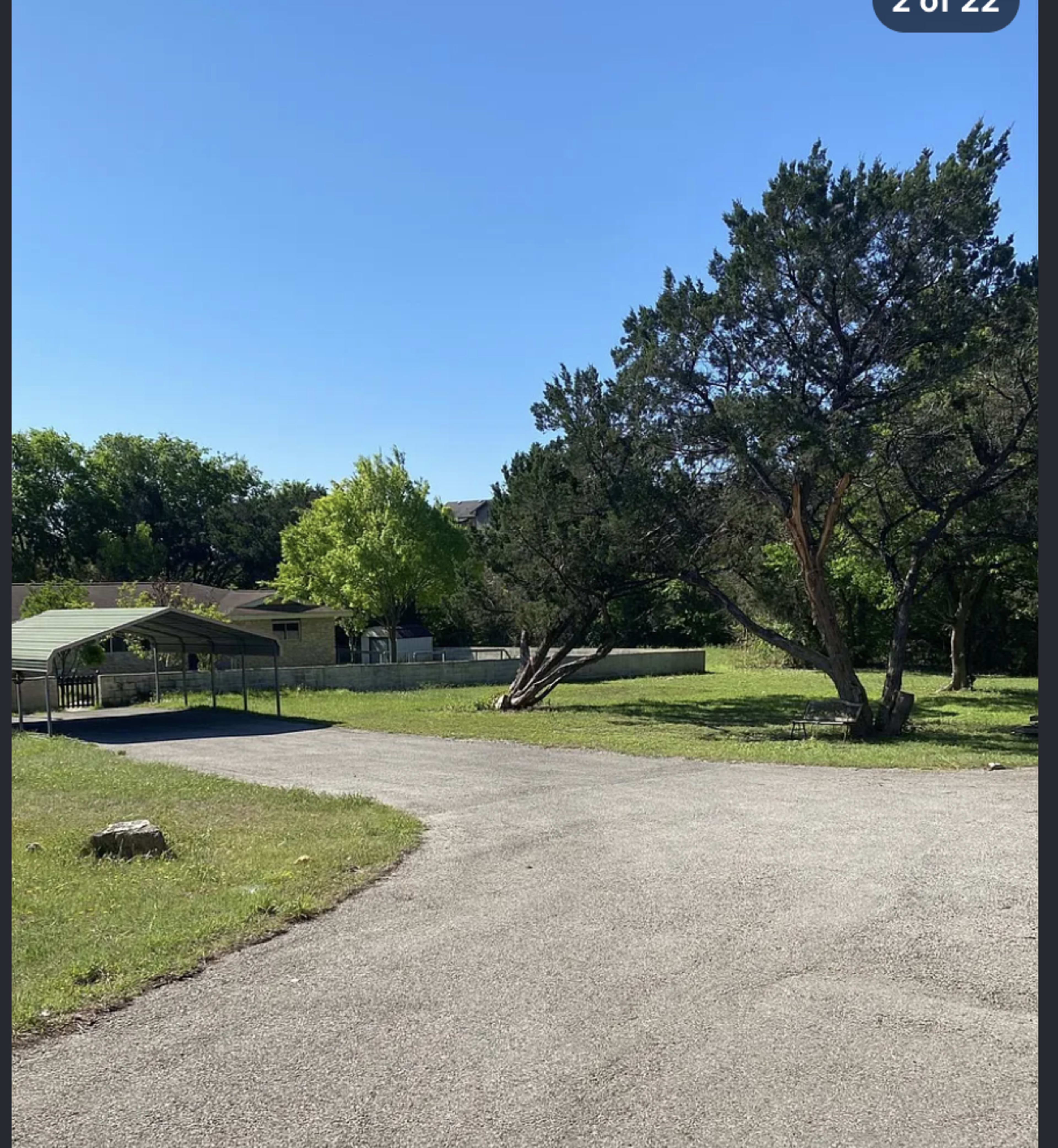 A winding gravel path leads through a grassy area with a few trees and a house in the background under a clear blue sky.