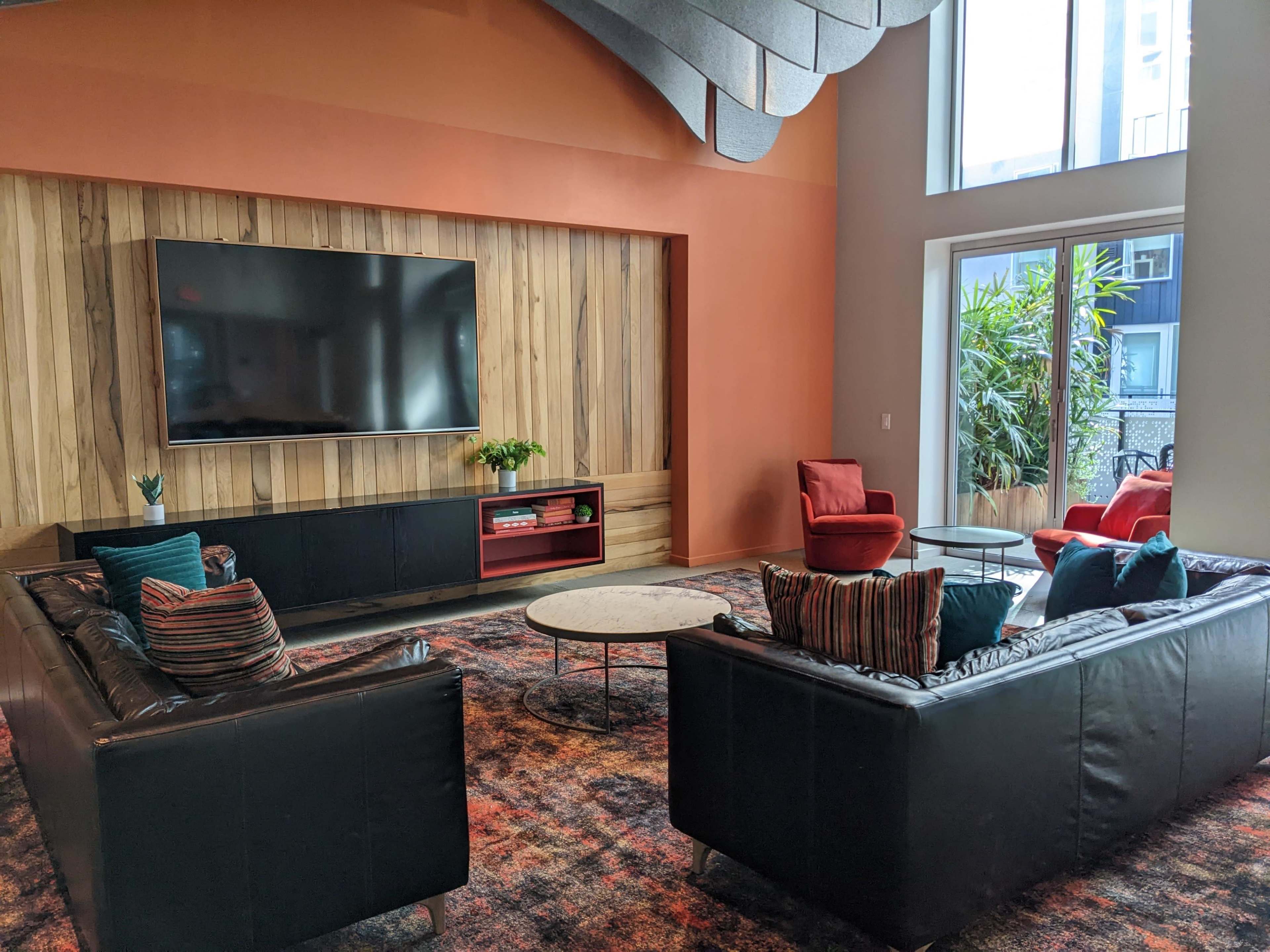 A modern lounge area with two black sofas, a round coffee table, and a large wall-mounted television against a backdrop of wooden paneling and orange walls.