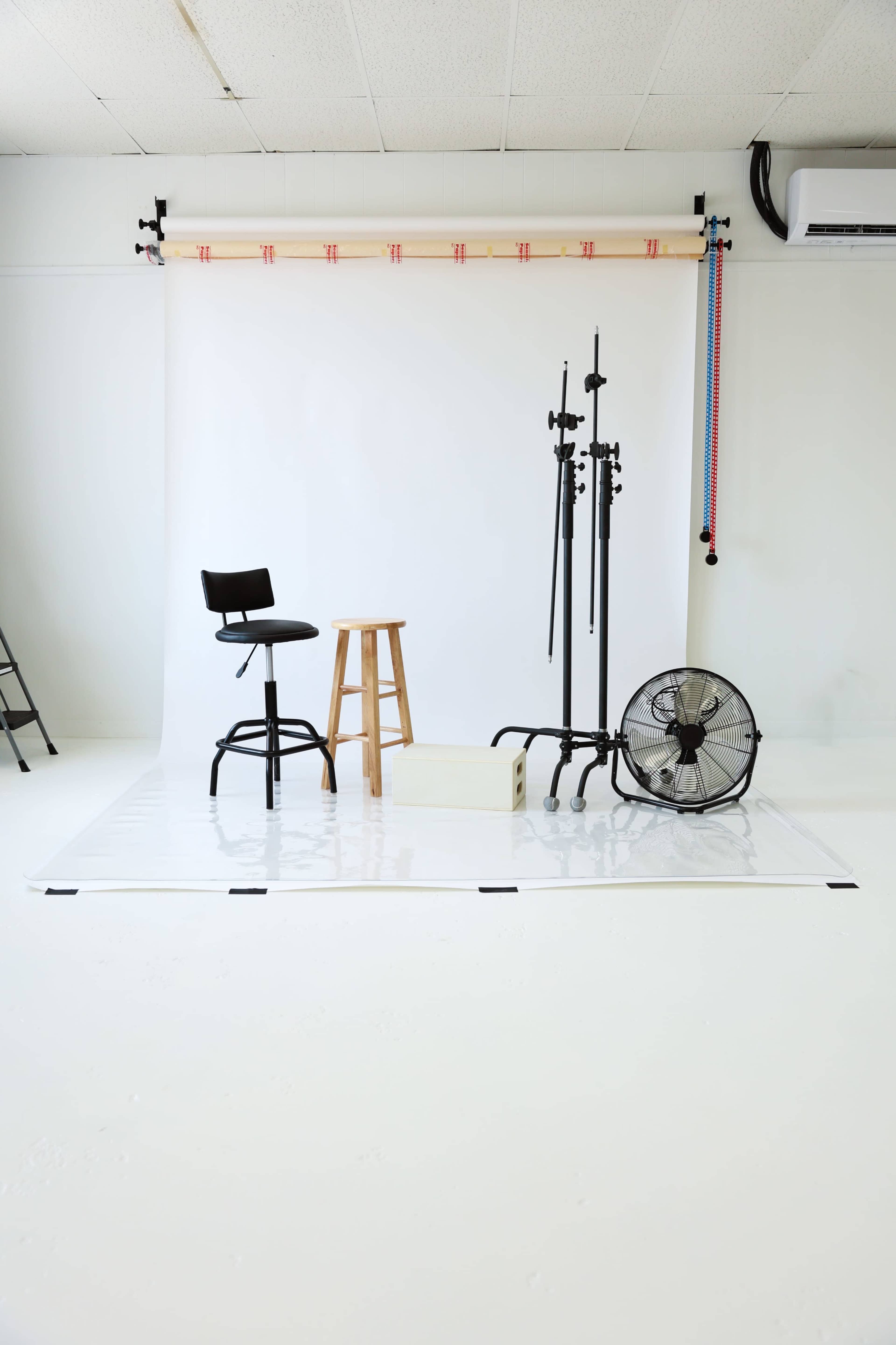 The image shows a minimalistic studio setup featuring a black stool, a wooden stool, lighting stands, a box, and a fan against a plain white backdrop.