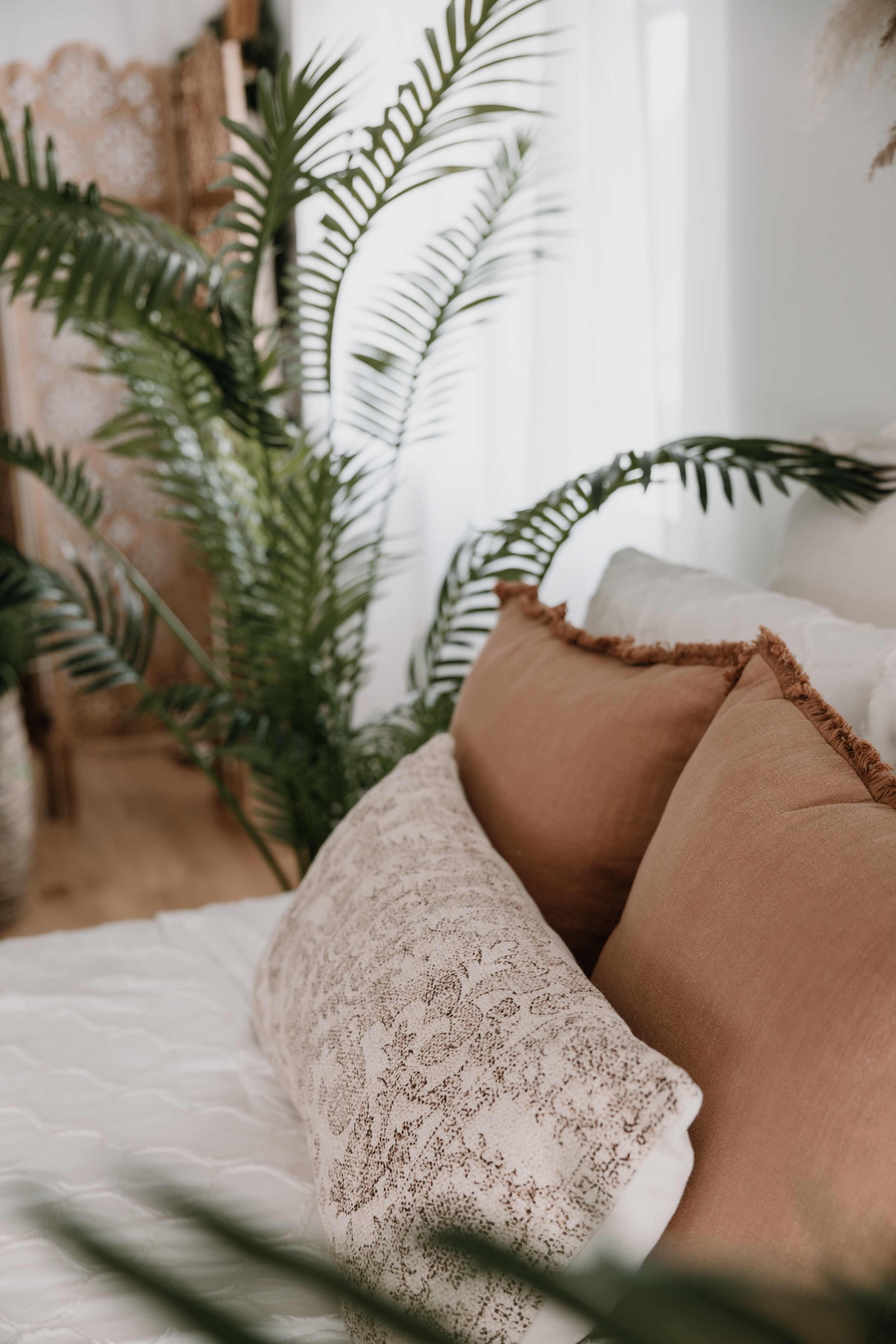 The image shows a cozy bed adorned with decorative pillows and surrounded by greenery in a bright, minimalist room.