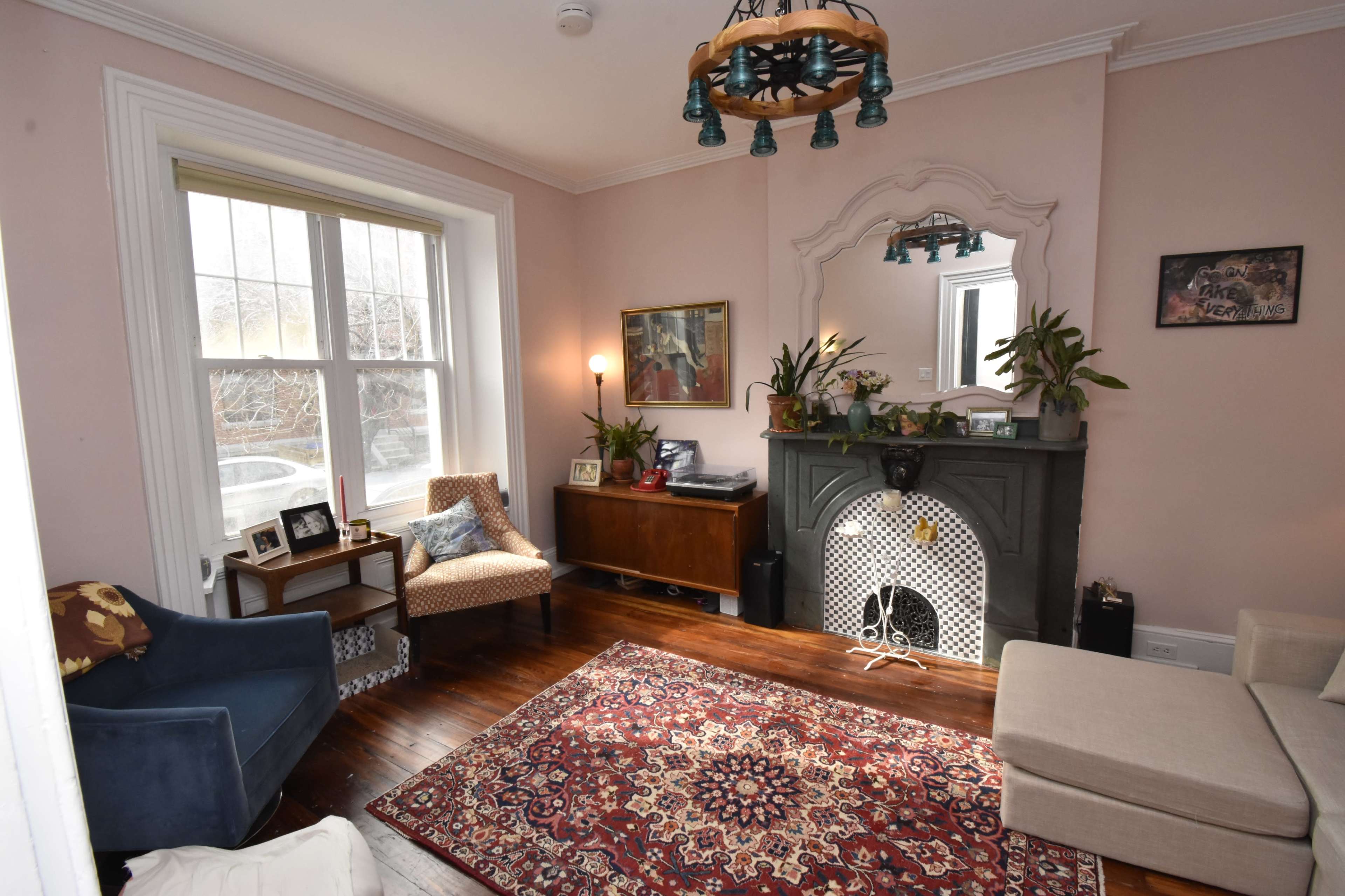 The image shows a cozy living room featuring a dark green fireplace, a beige sofa, a blue armchair, and a patterned rug on wooden floors.