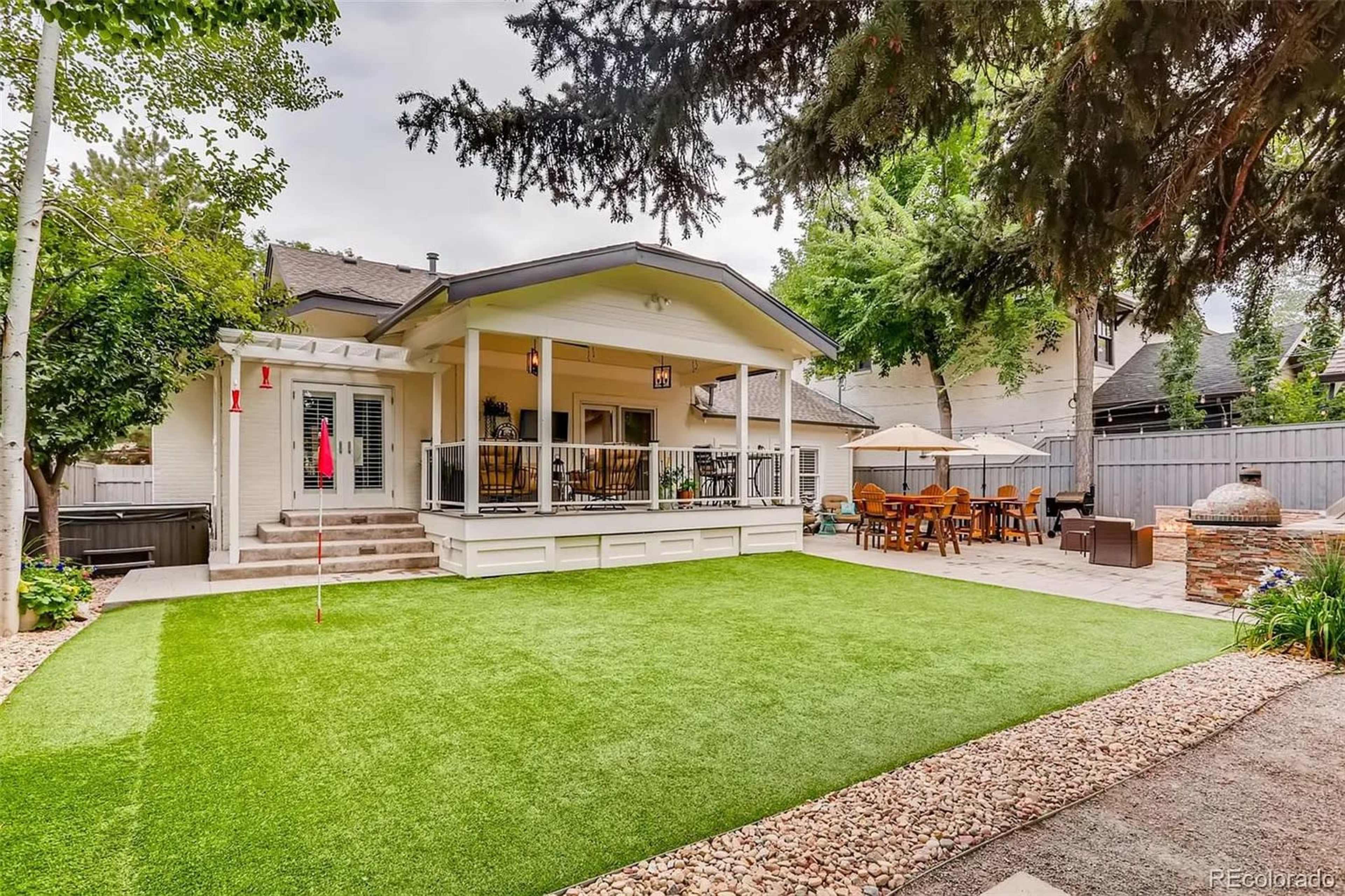 The image shows a backyard with a putting green, a covered patio area with dining furniture, and landscaping that includes trees and decorative rocks.