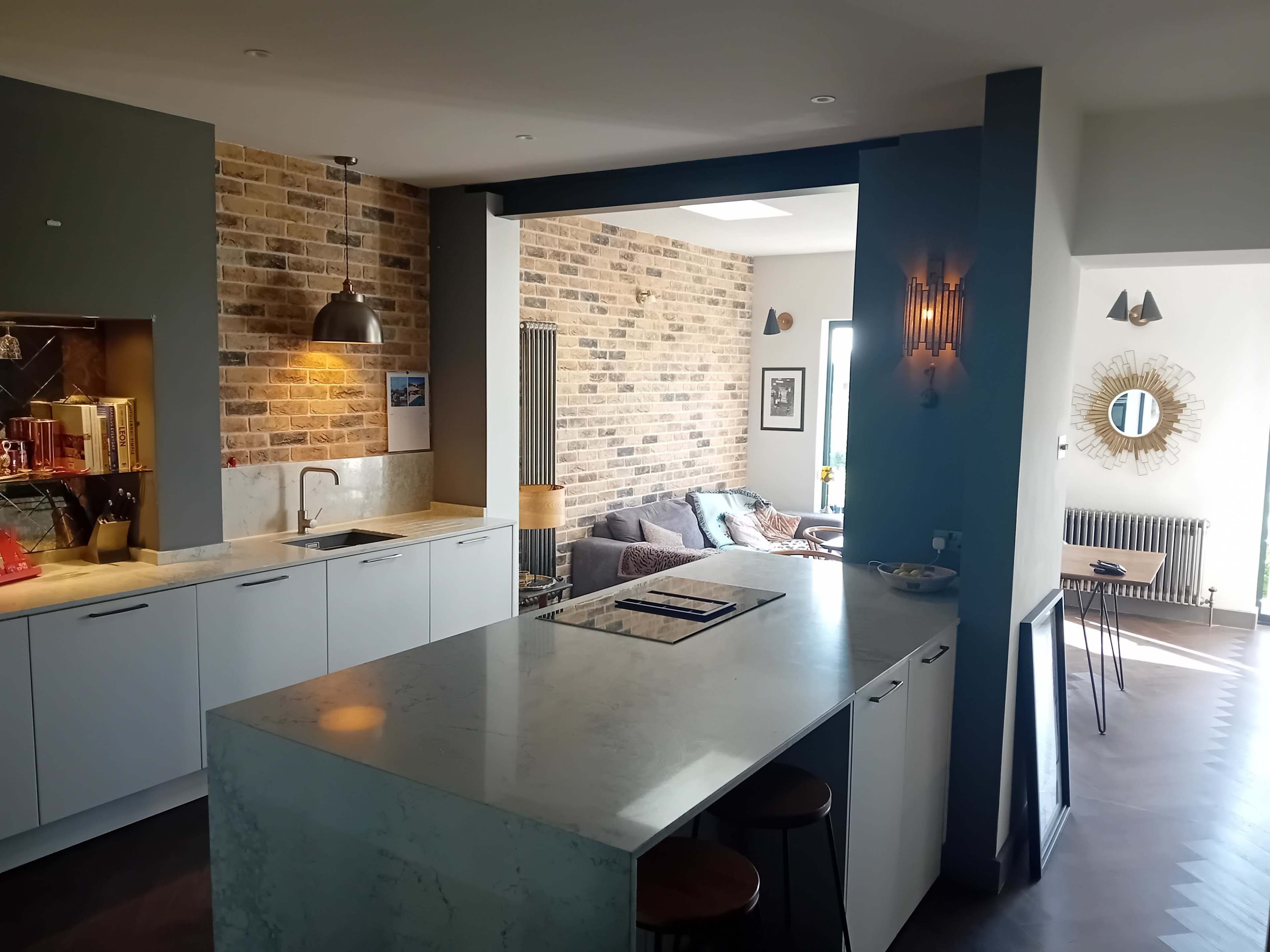 The image shows a modern, open-concept kitchen and living area featuring a marble kitchen island, exposed brick walls, and a comfortable seating arrangement in the living space.