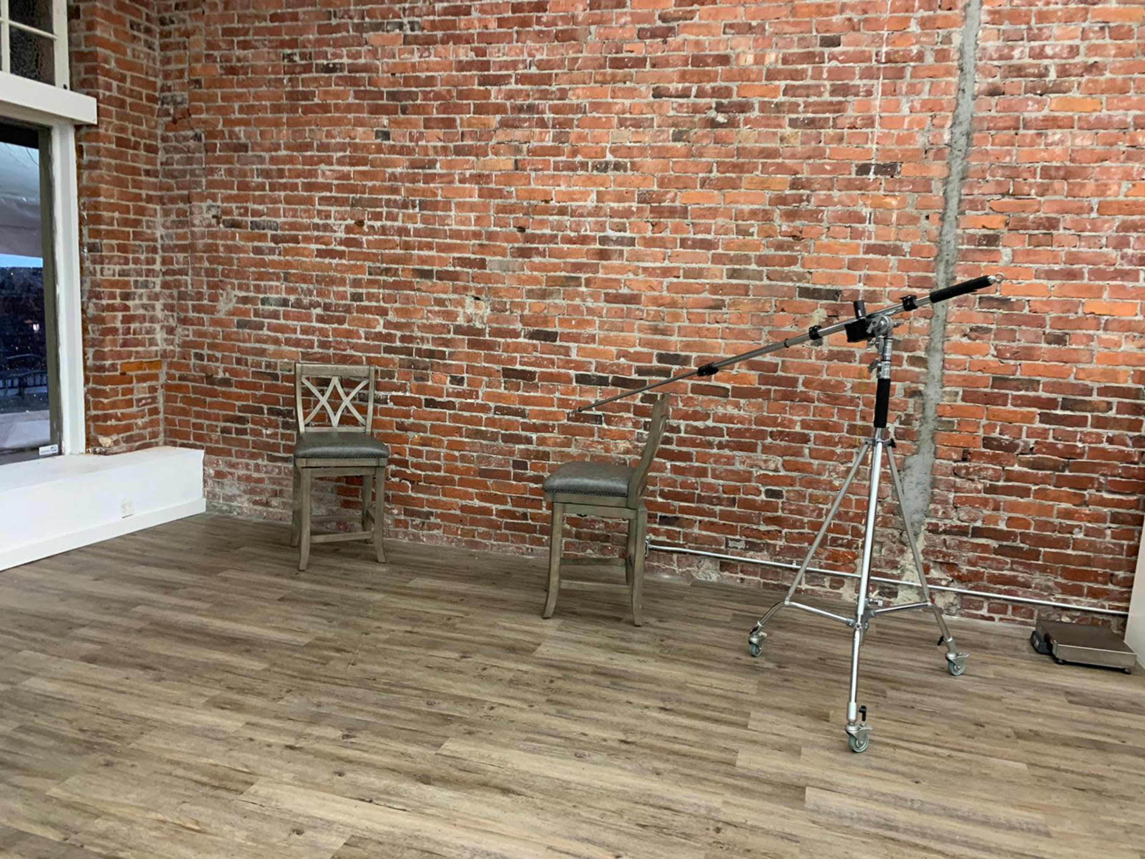 A room with exposed brick walls, two wooden chairs, and a camera tripod positioned nearby on a wooden floor.