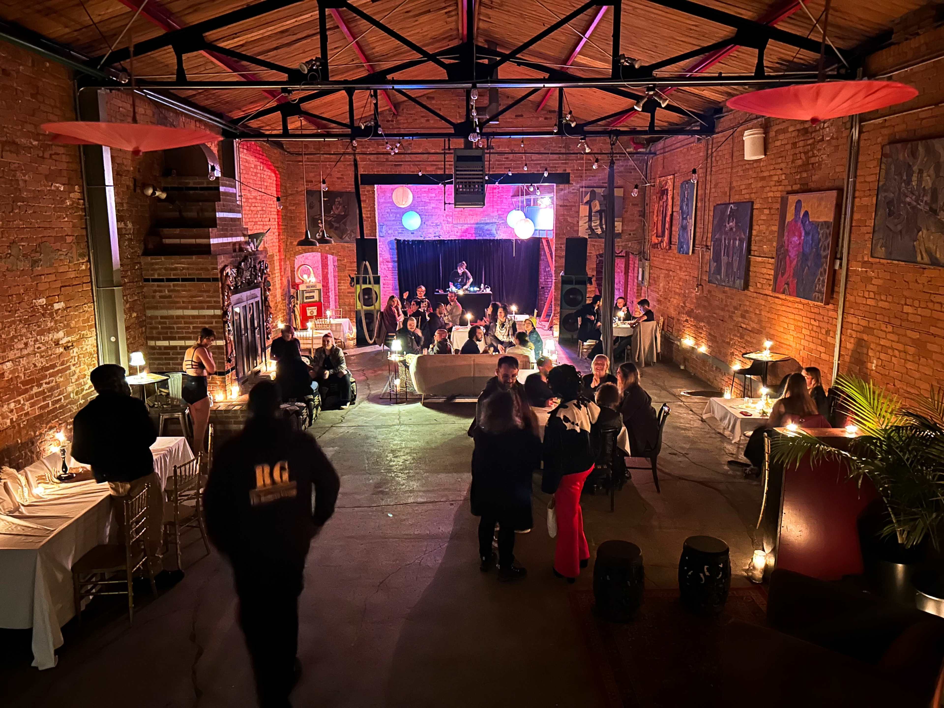 A lively event takes place in a spacious brick-walled venue, featuring guests seated at tables and a performance stage illuminated with colorful lights.