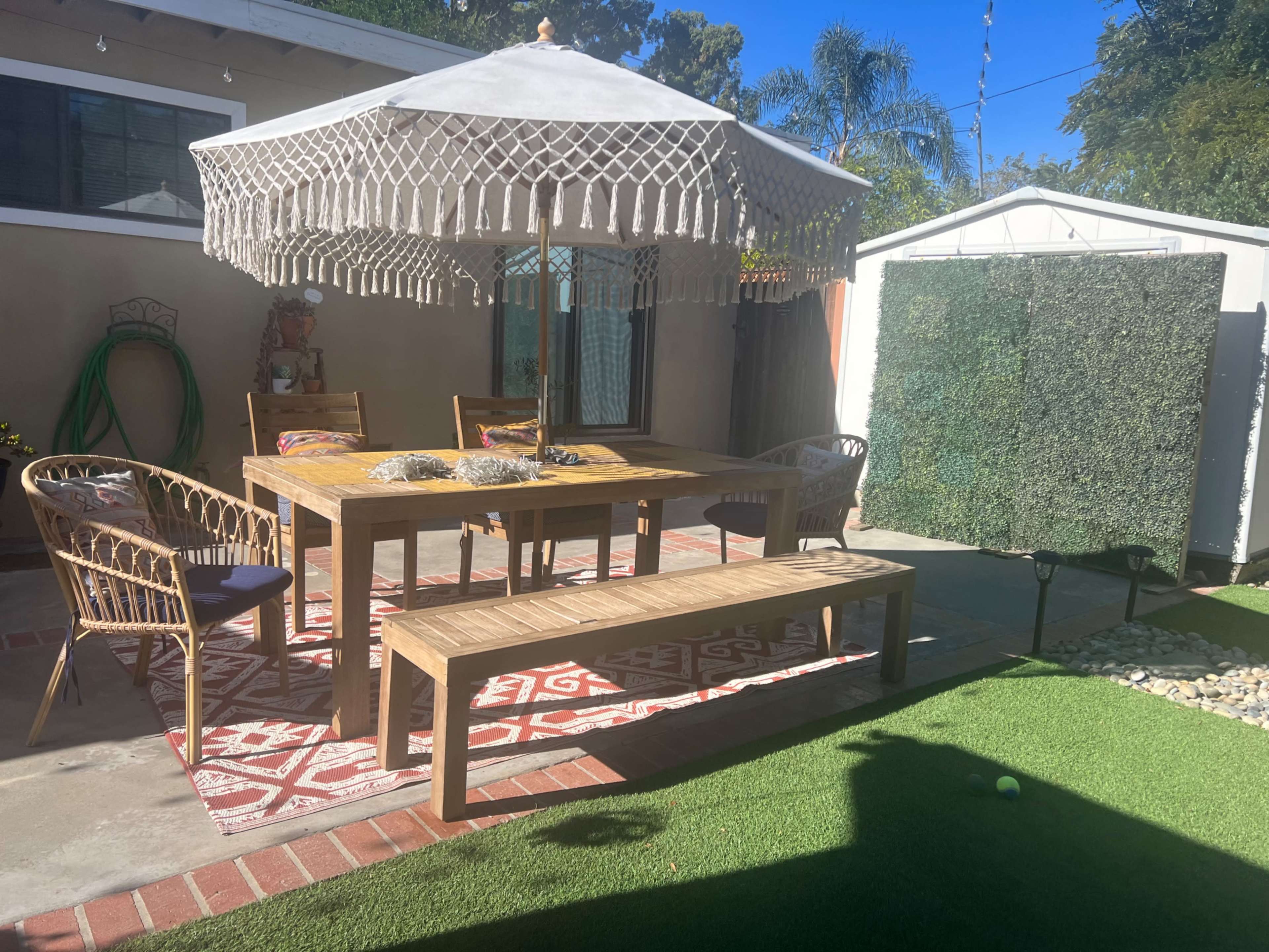 The image shows a shaded outdoor dining area with a wooden table, wicker chairs, and a decorative parasol, surrounded by greenery and a storage shed in the background.