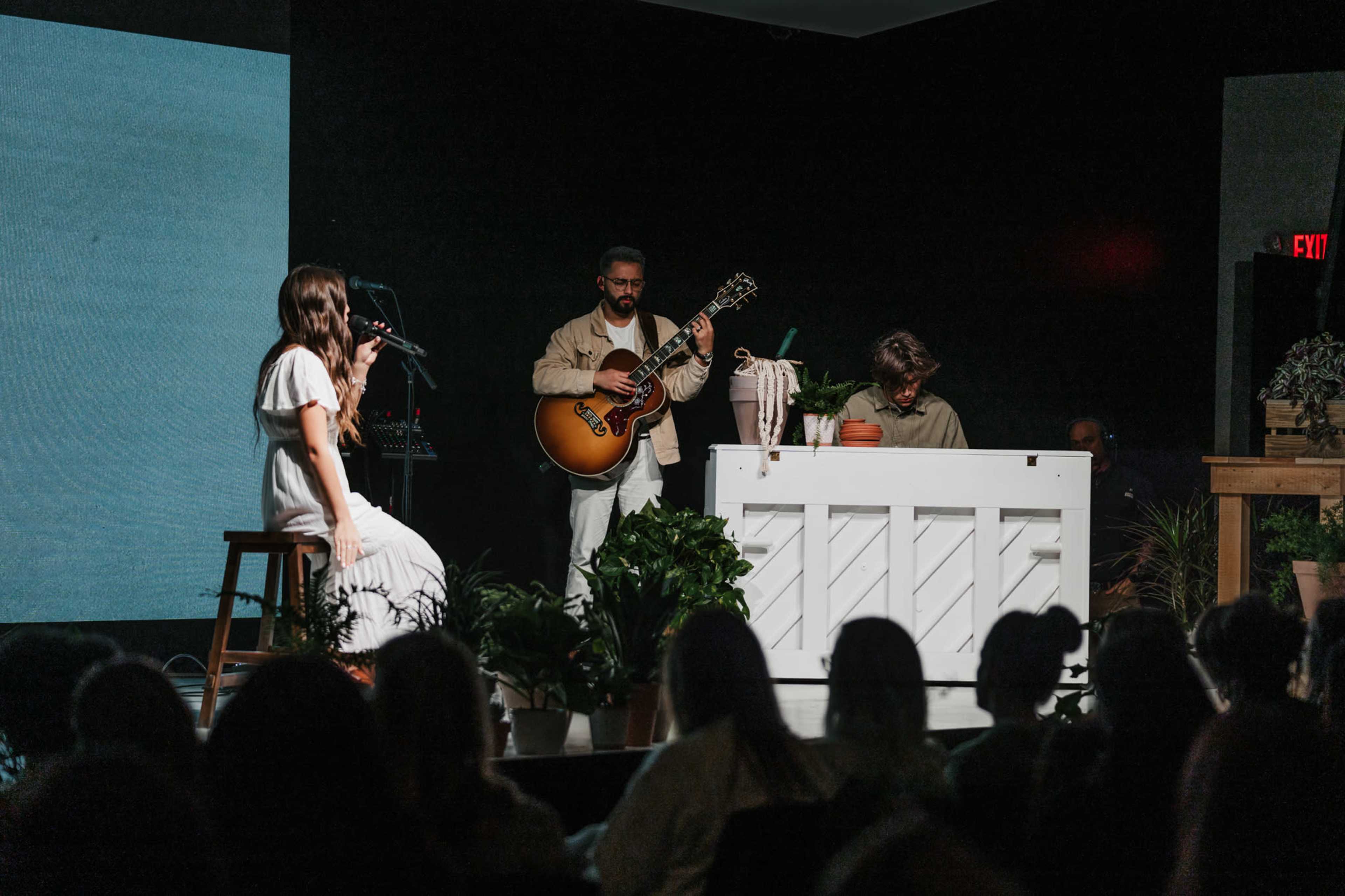 A musician performs on stage with a guitar while another musician plays a piano, accompanied by a vocalist sitting on a stool.