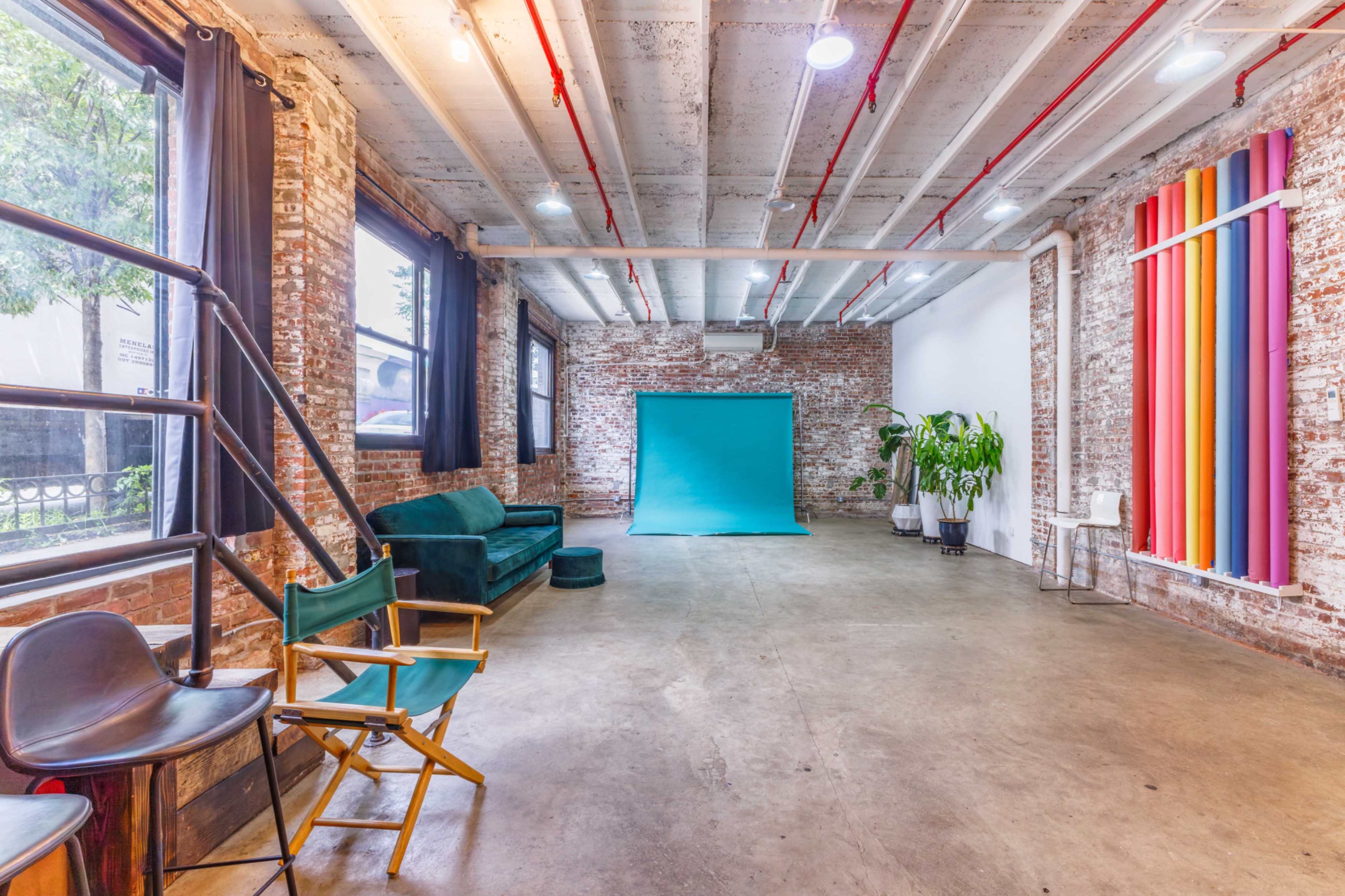 The image shows a spacious, industrial-style room with exposed brick walls, concrete flooring, a turquoise backdrop, and seating options including a green sofa and director's chairs.