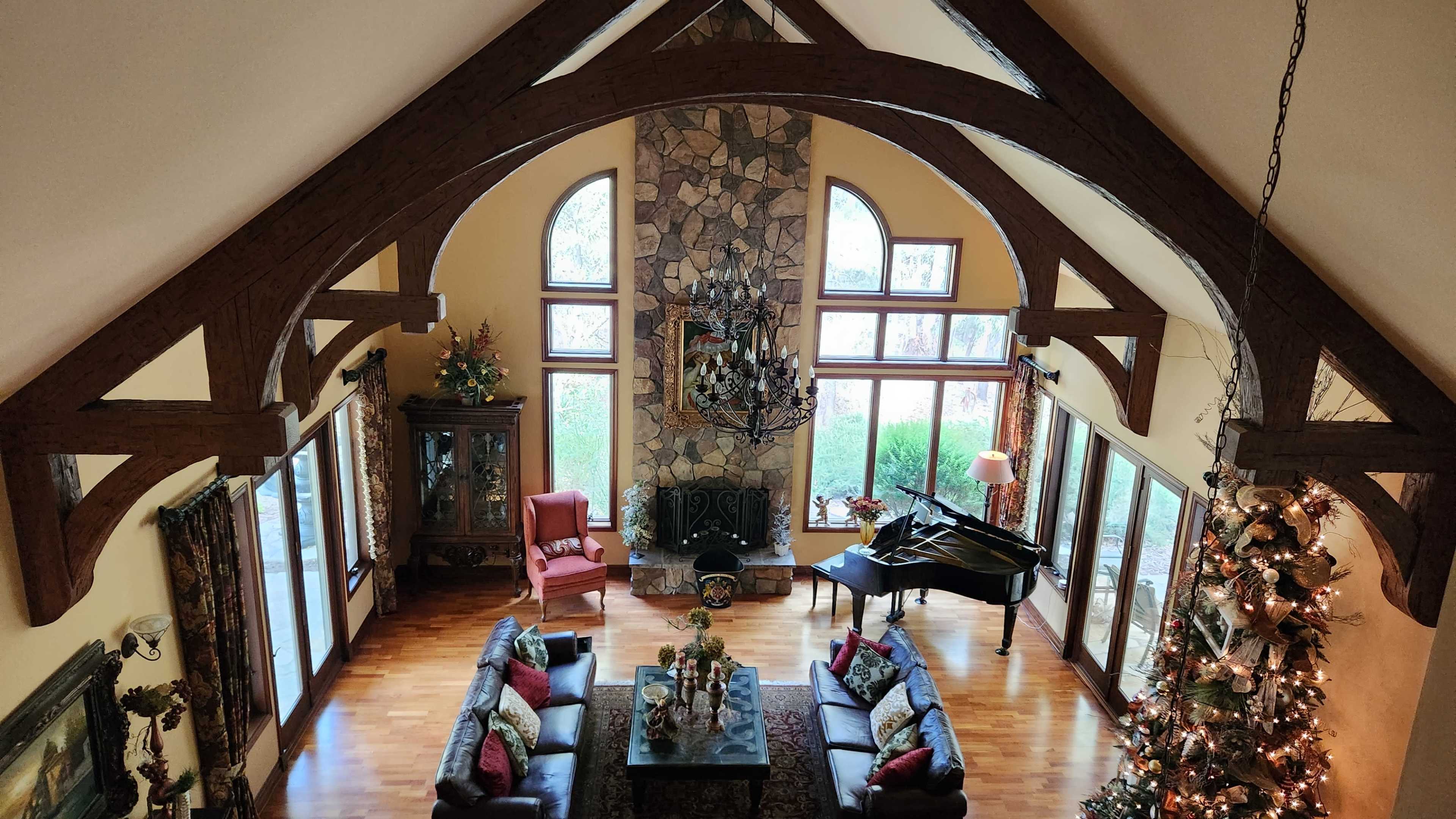 The image shows a spacious living room with high ceilings, wooden beams, a grand piano, and large windows overlooking a garden, decorated for the holidays.