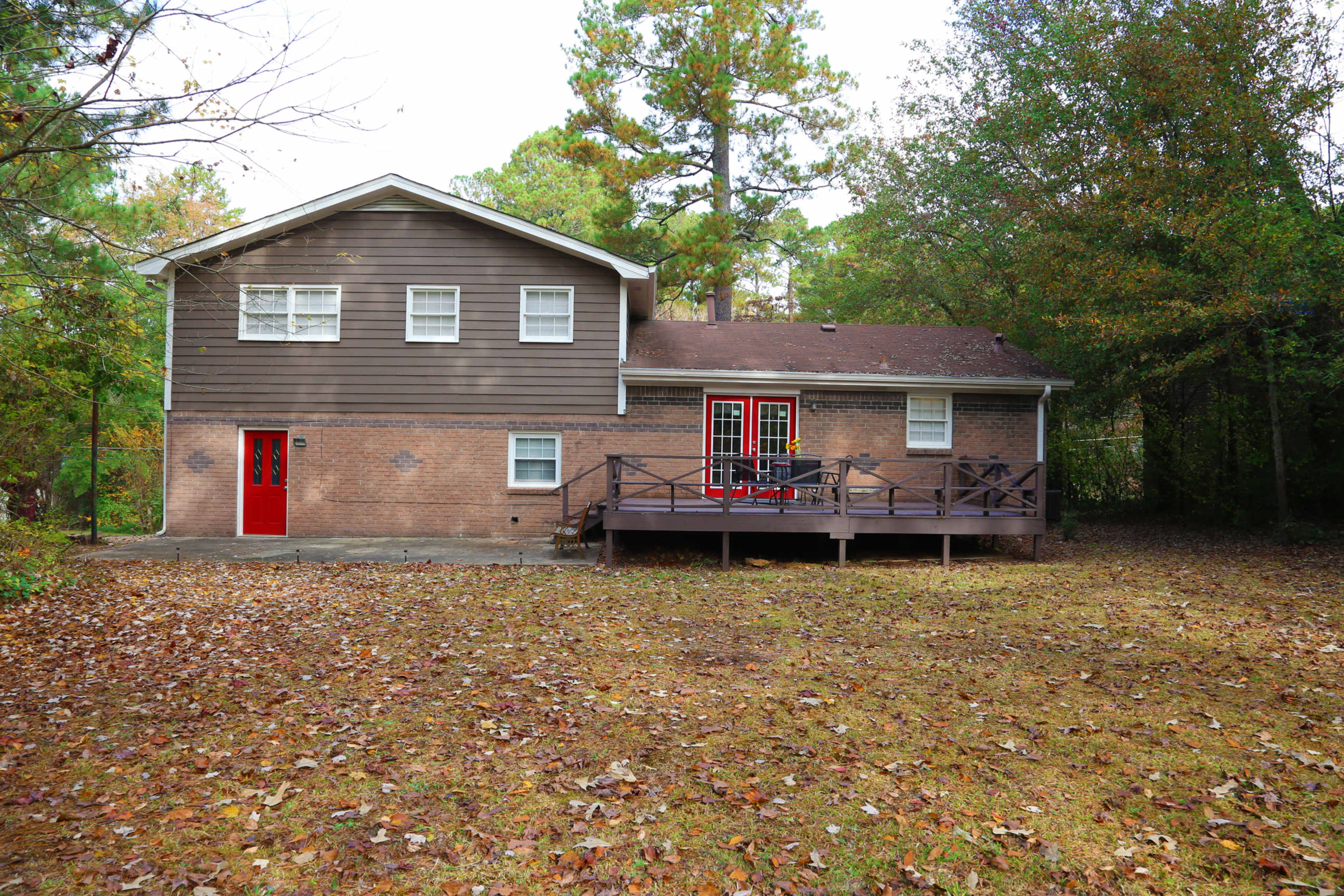 A two-story brown house with red doors sits on a grassy yard surrounded by trees.