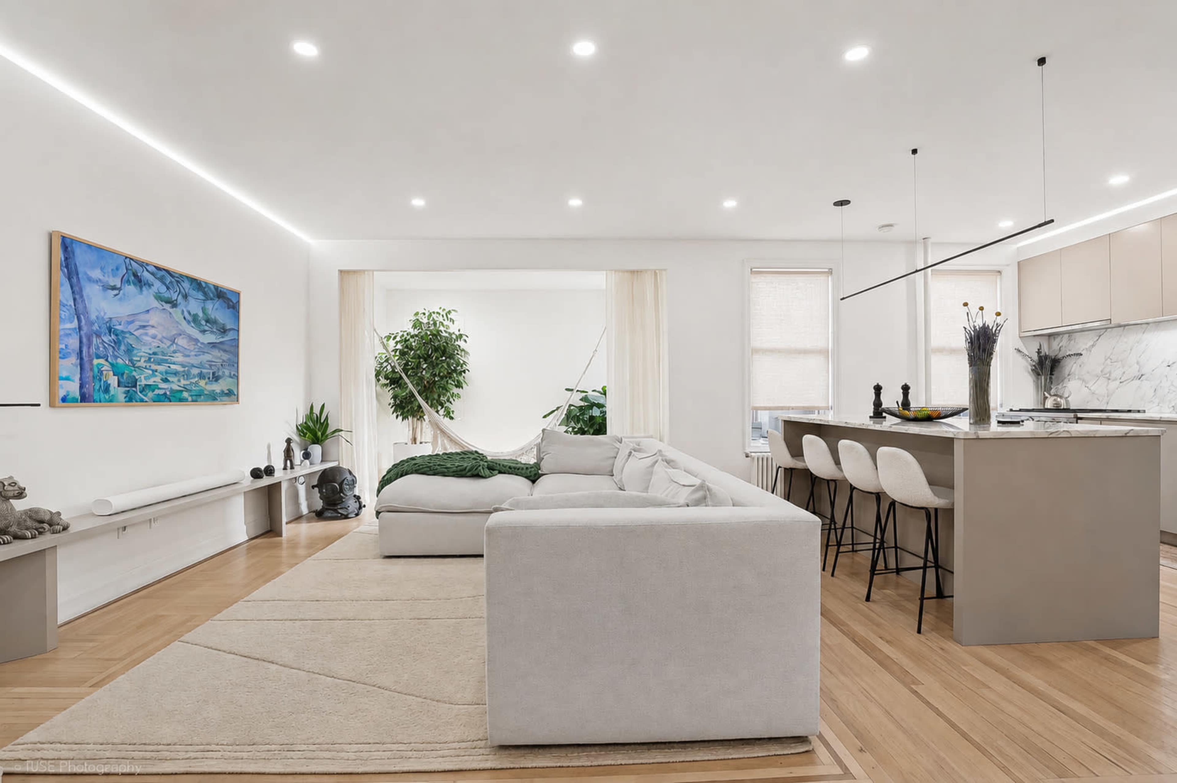 The image shows a modern living room and kitchen area featuring a light-colored sectional sofa, a marble counter with bar stools, and a large artwork on the wall.