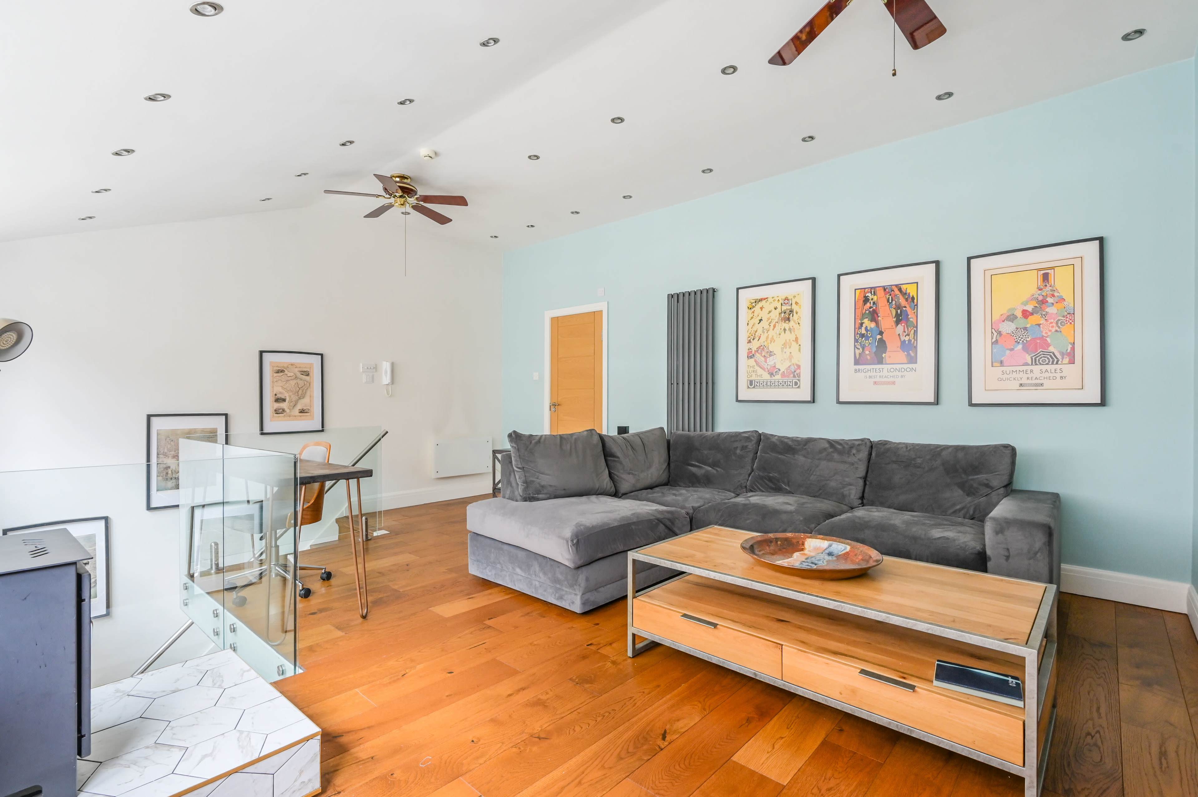 The image shows a modern living room with hardwood flooring, a large gray sectional sofa, and a glass partition near a workspace with a desk and chair.