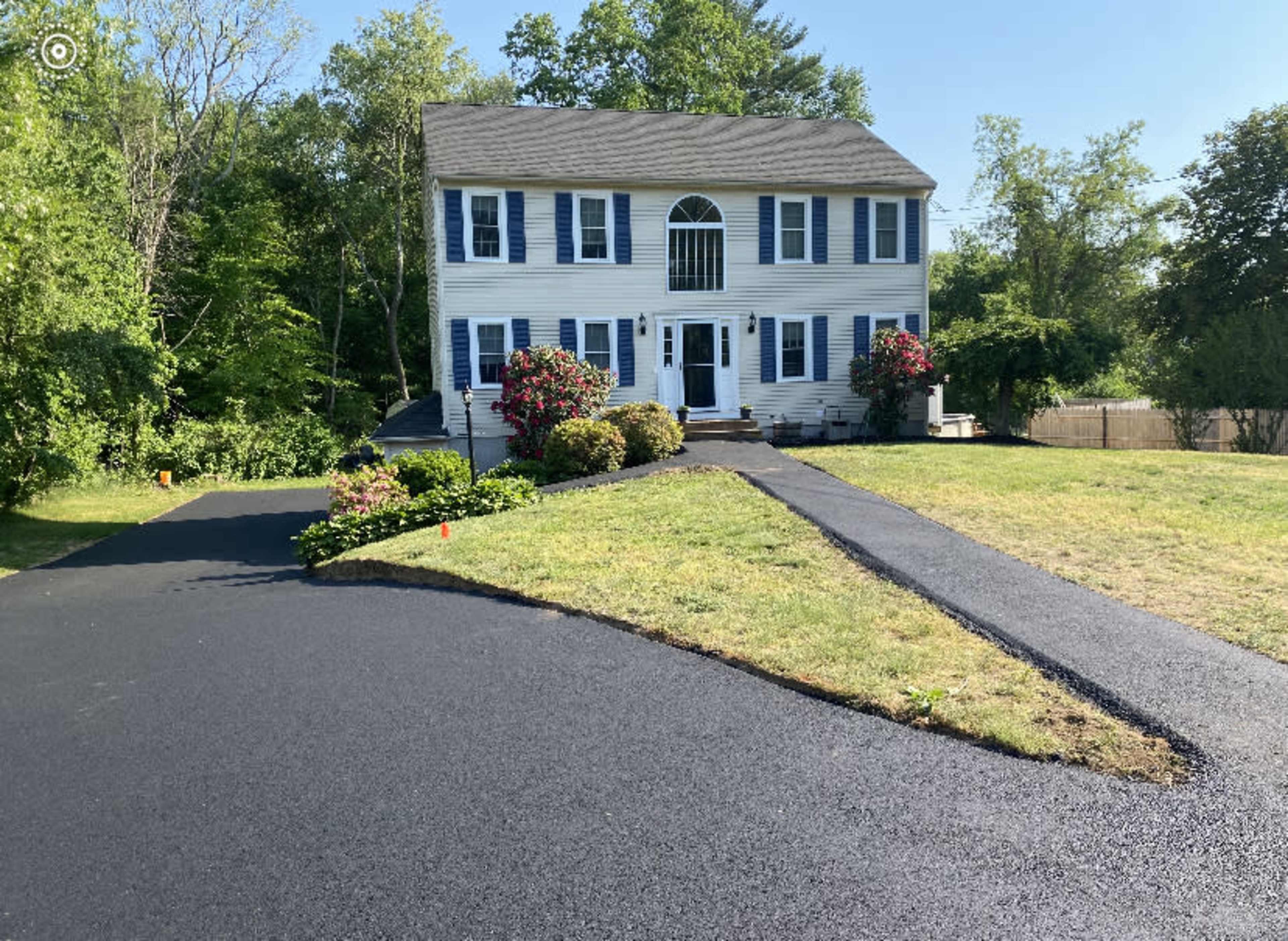 A two-story blue and white house with a front porch and landscaped yard sits on a newly asphalted driveway.