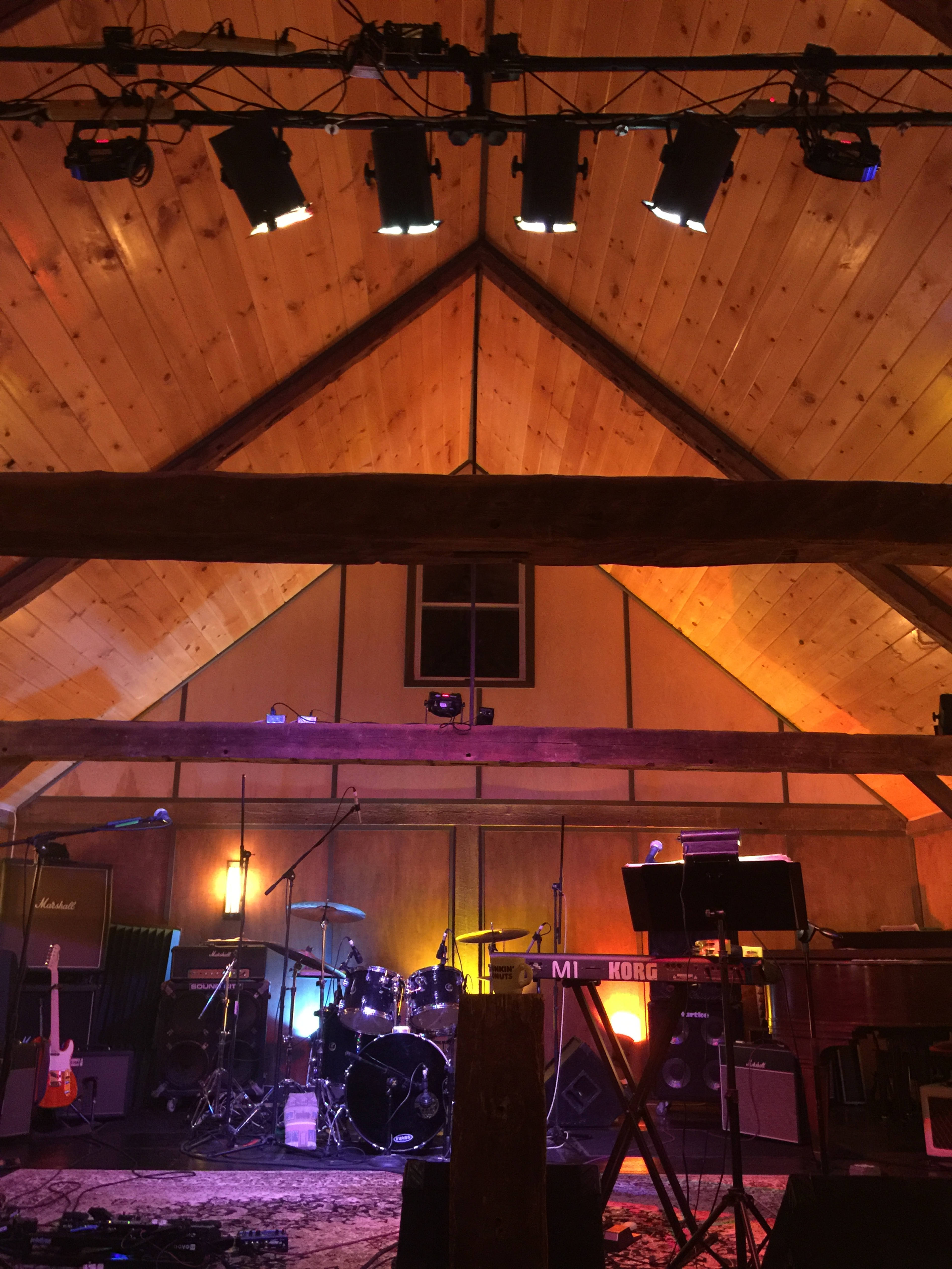 A music performance space with wooden beams, stage lighting, and various musical instruments set up, including drums and keyboards.