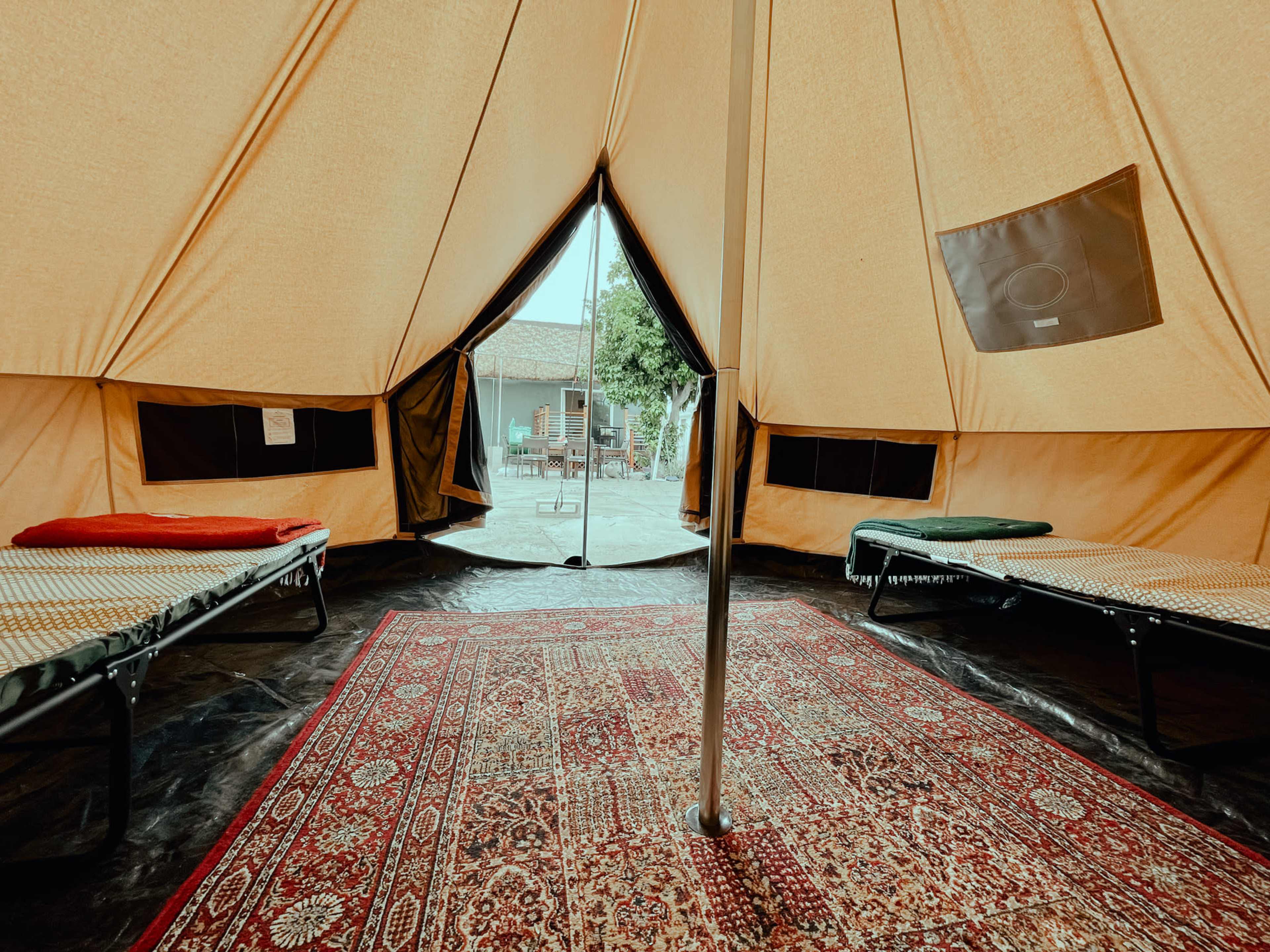 The interior of a large tent features two cots on either side and a decorative rug in the center, with an opening leading to an outdoor area.