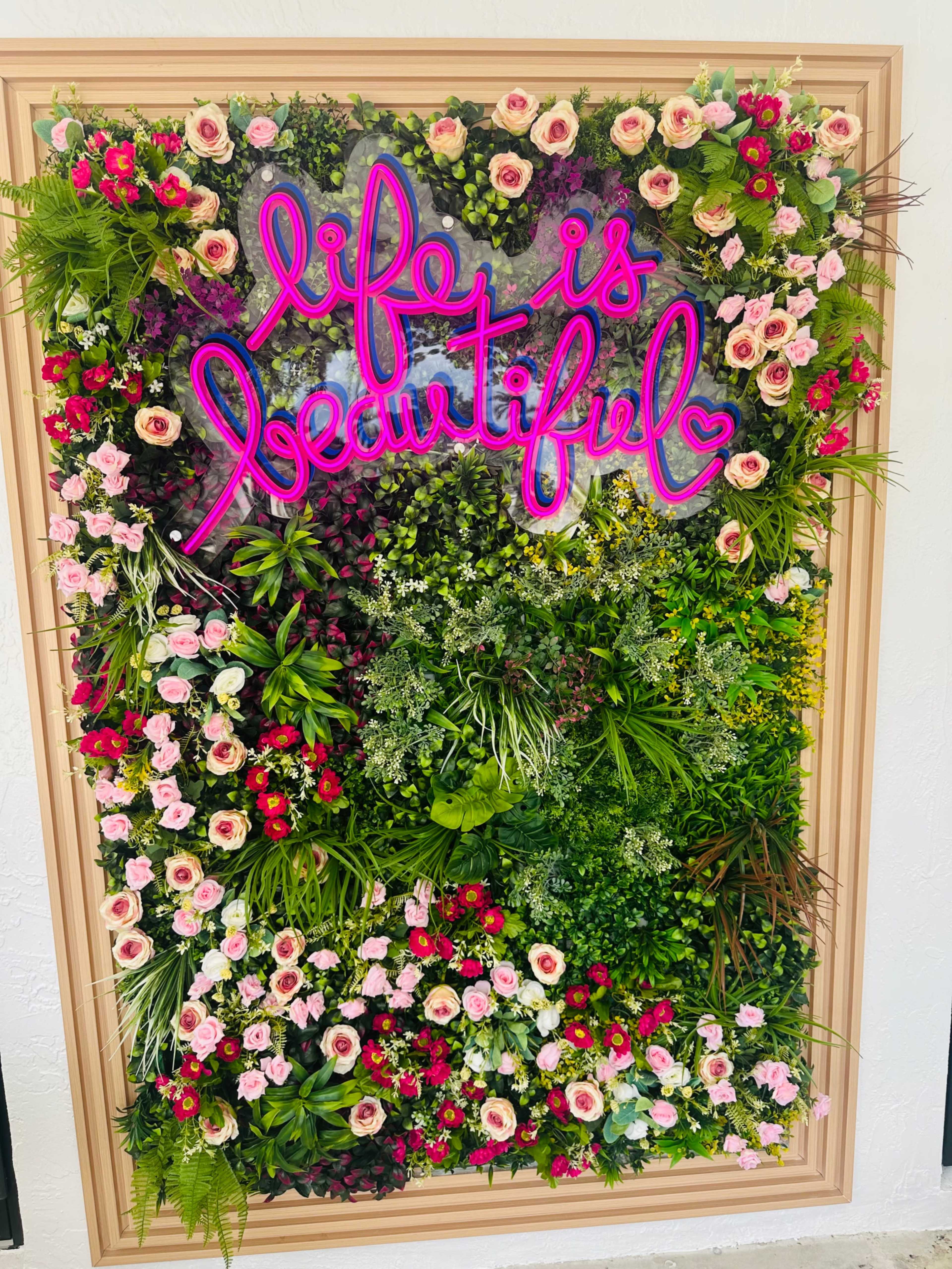 A colorful floral wall displays the neon phrase "life is beautiful" surrounded by various flowers and greenery.