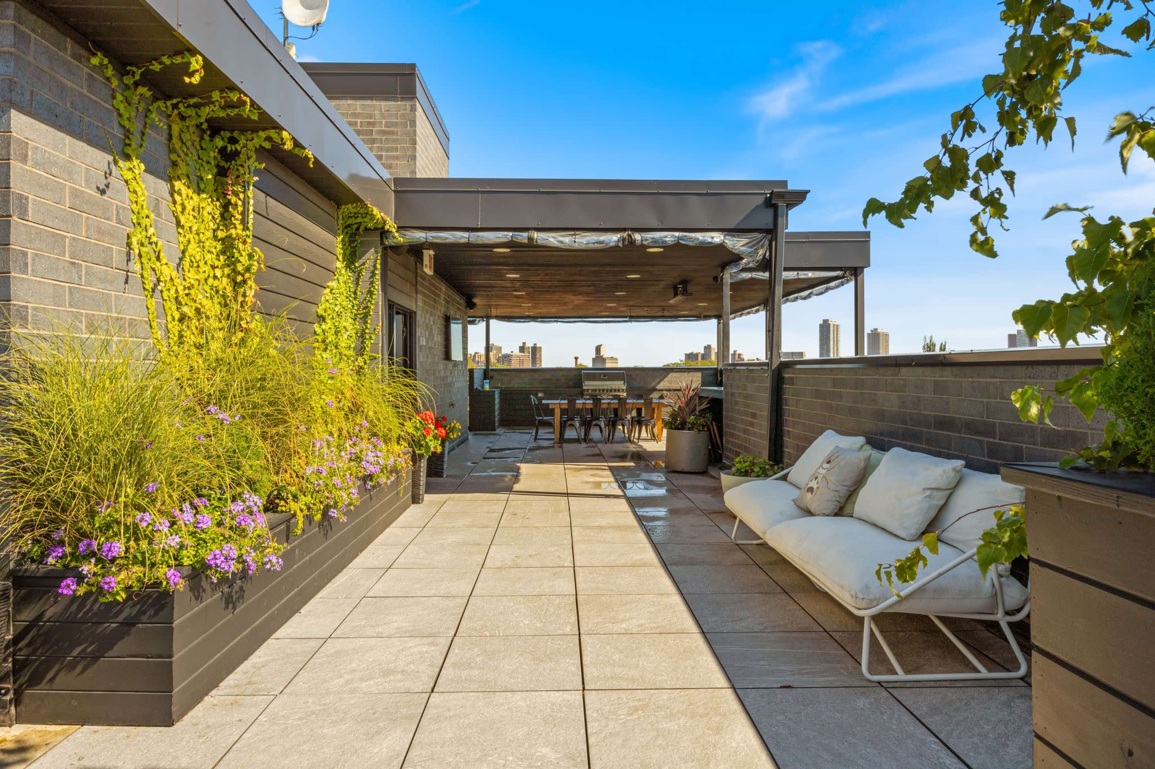 The image shows a rooftop terrace with potted plants, seating, and a dining area overlooking a city skyline.