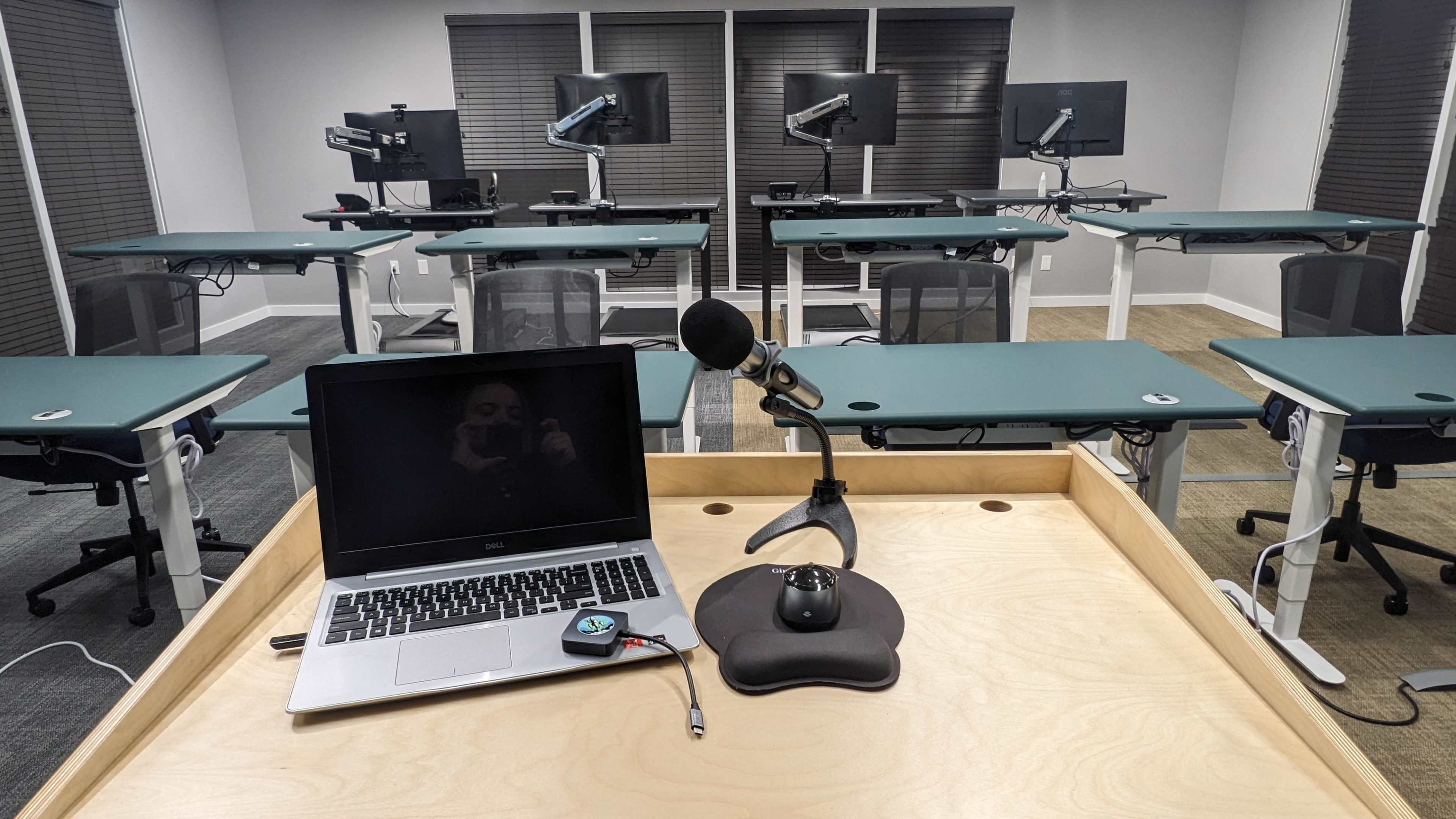 A desktop setup with a laptop and microphone is positioned at the front of a classroom filled with adjustable desks and computer monitors.