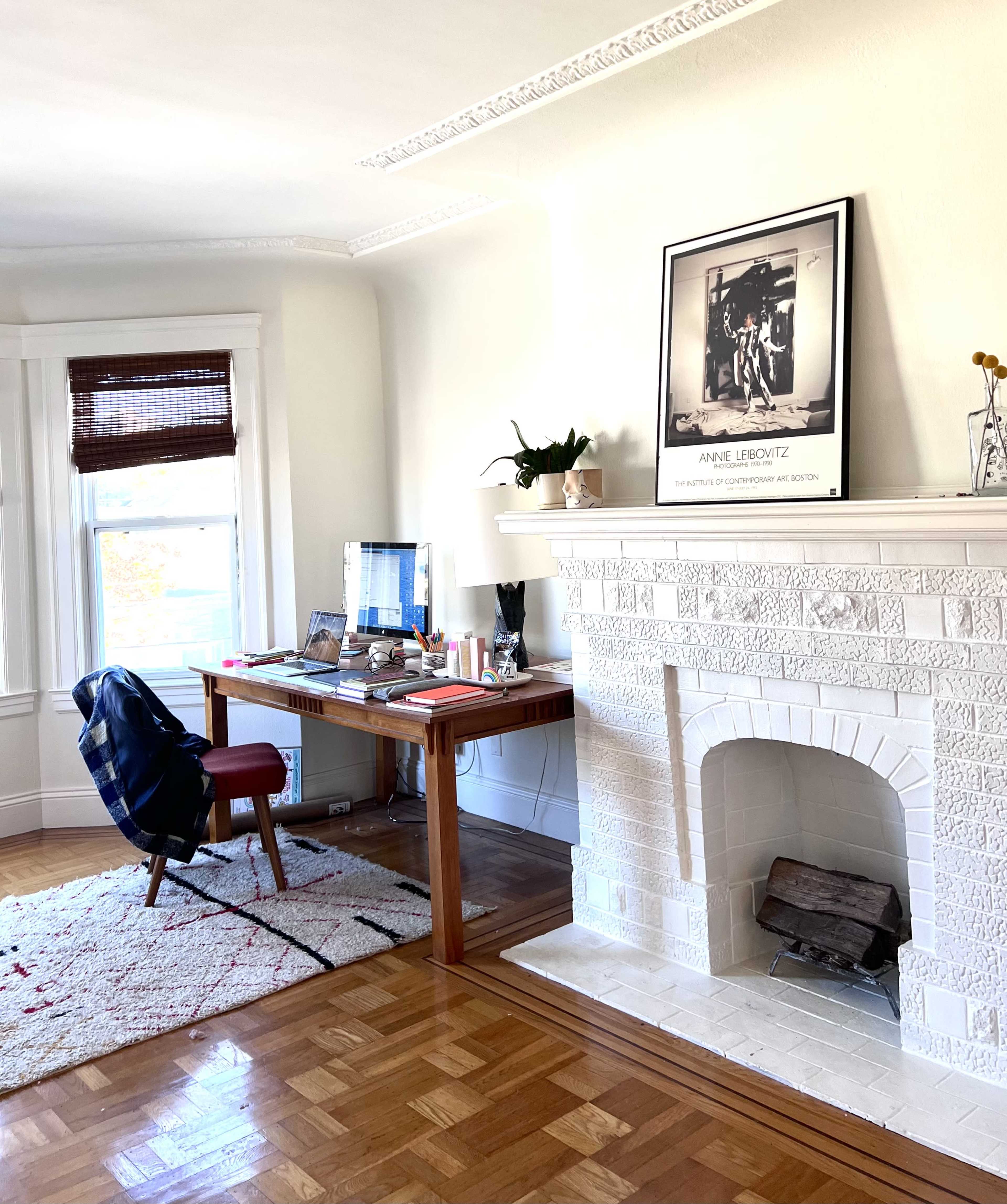 A home office with a wooden desk, a chair, a computer setup, and a fireplace adorned with a framed photograph and plants.