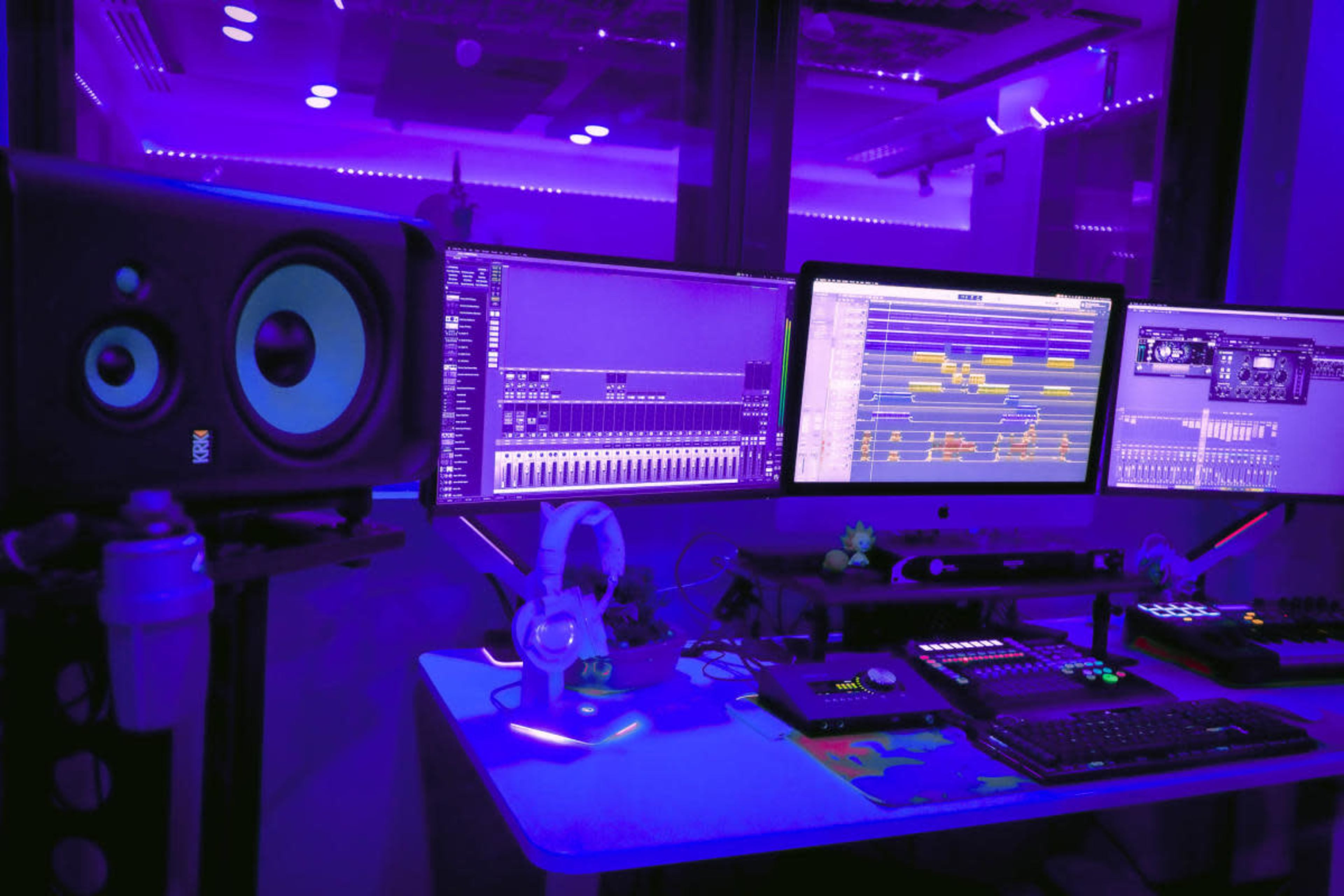 A music production studio features dual computer monitors displaying audio software, surrounded by various equipment, with purple lighting illuminating the space.