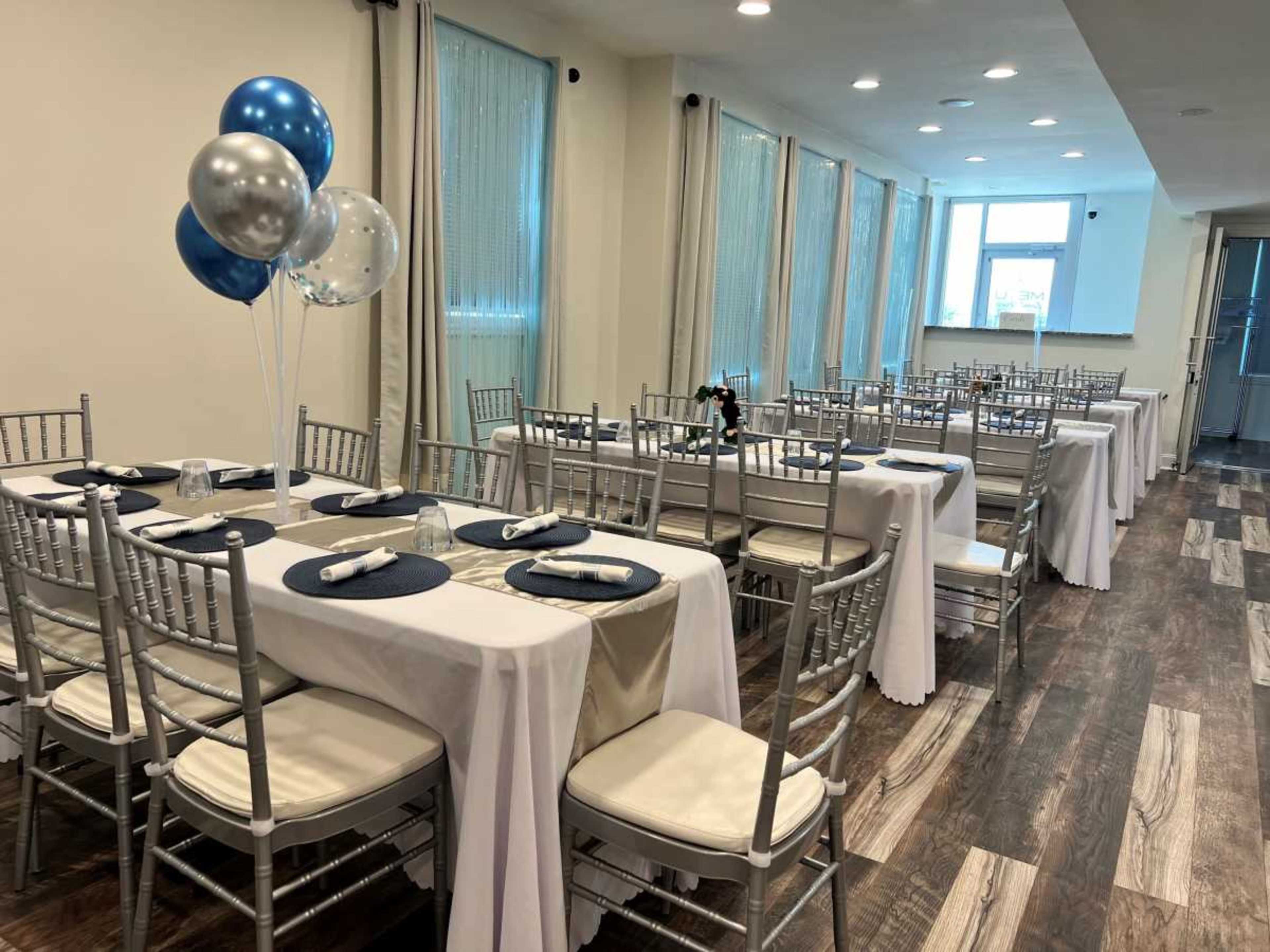 The image shows a set-up for an event in a banquet room with silver and blue balloons and neatly arranged tables covered in white tablecloths.
