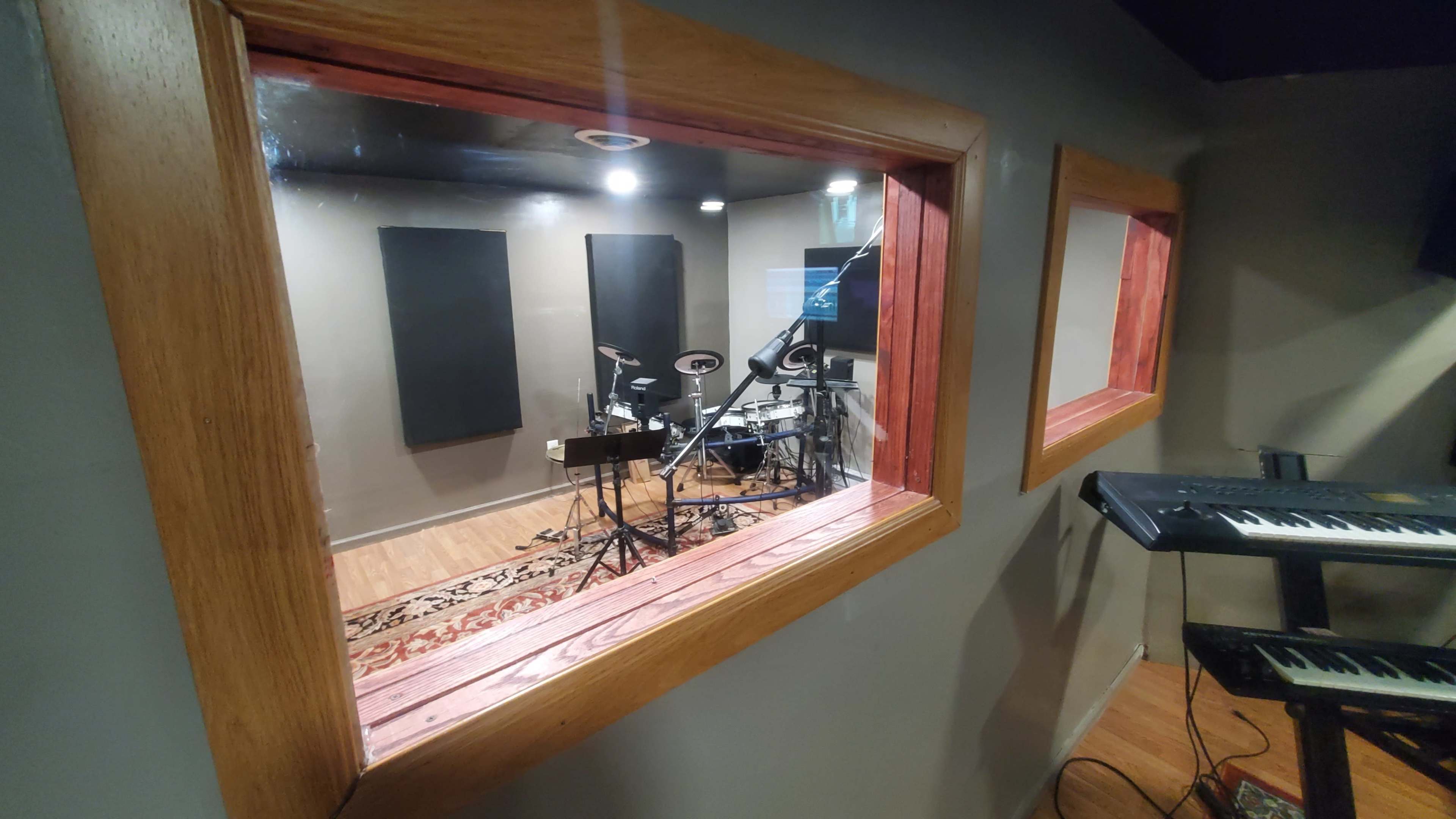 A recording studio room is visible through a set of glass windows, featuring a drum set and electronic keyboard poised for use.