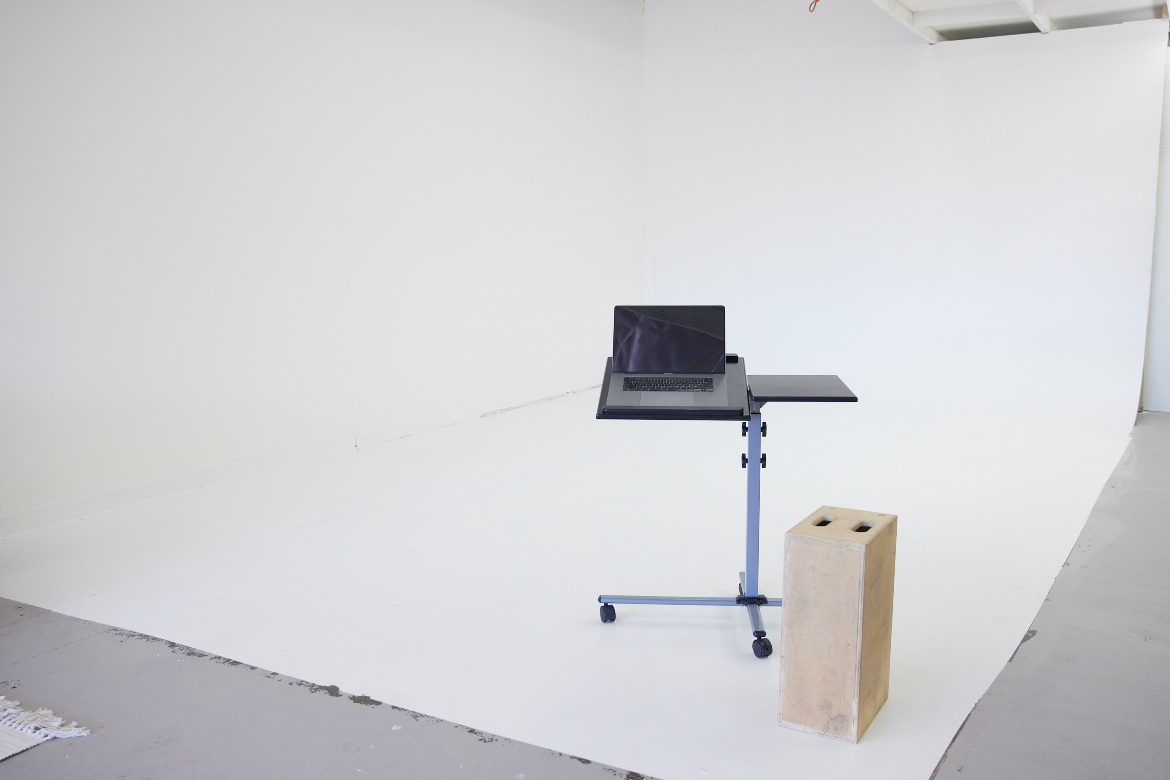 The image shows a minimalist, empty studio space featuring a laptop on a mobile stand next to a concrete block.
