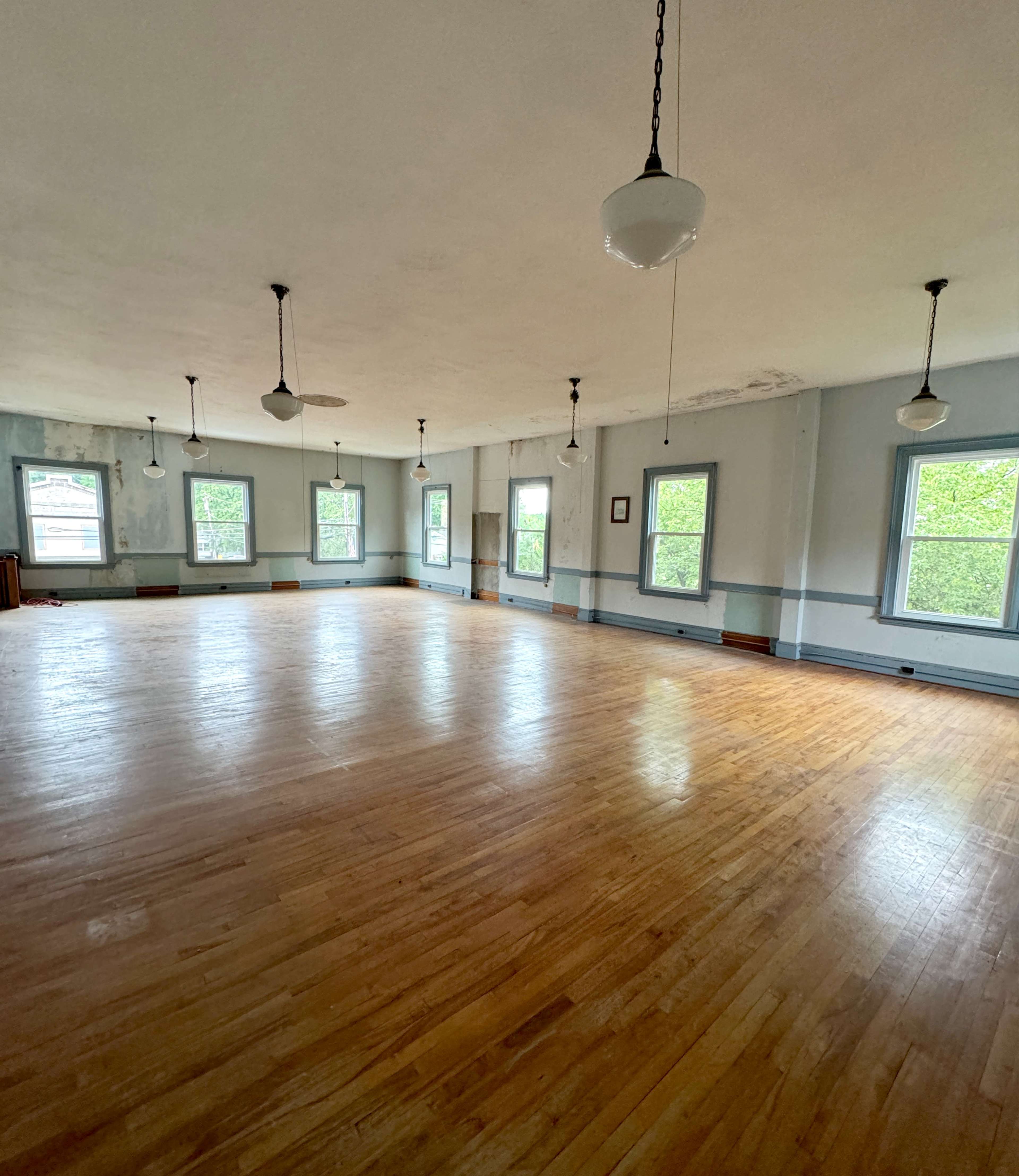 A spacious, empty room with hardwood floors and multiple windows, illuminated by hanging light fixtures.